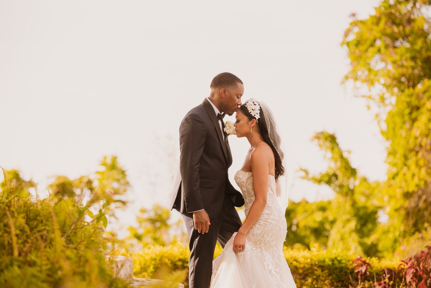 Newlyweds embrace in sunlit garden surrounded by blooming flowers and greenery.