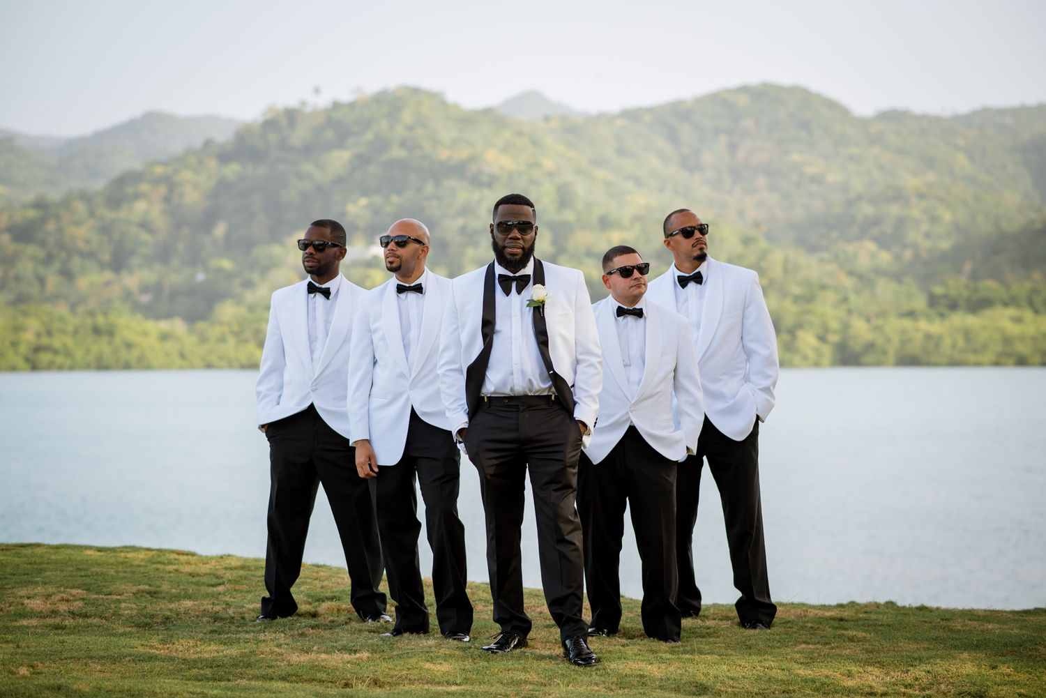 Groomsmen in formal white shirts and black pants pose by waterfront.