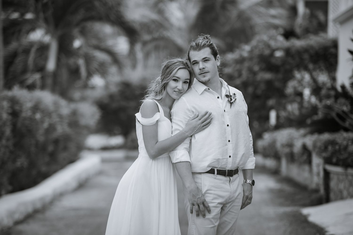 Black and white photo of newlyweds walking together on a tree-lined path.