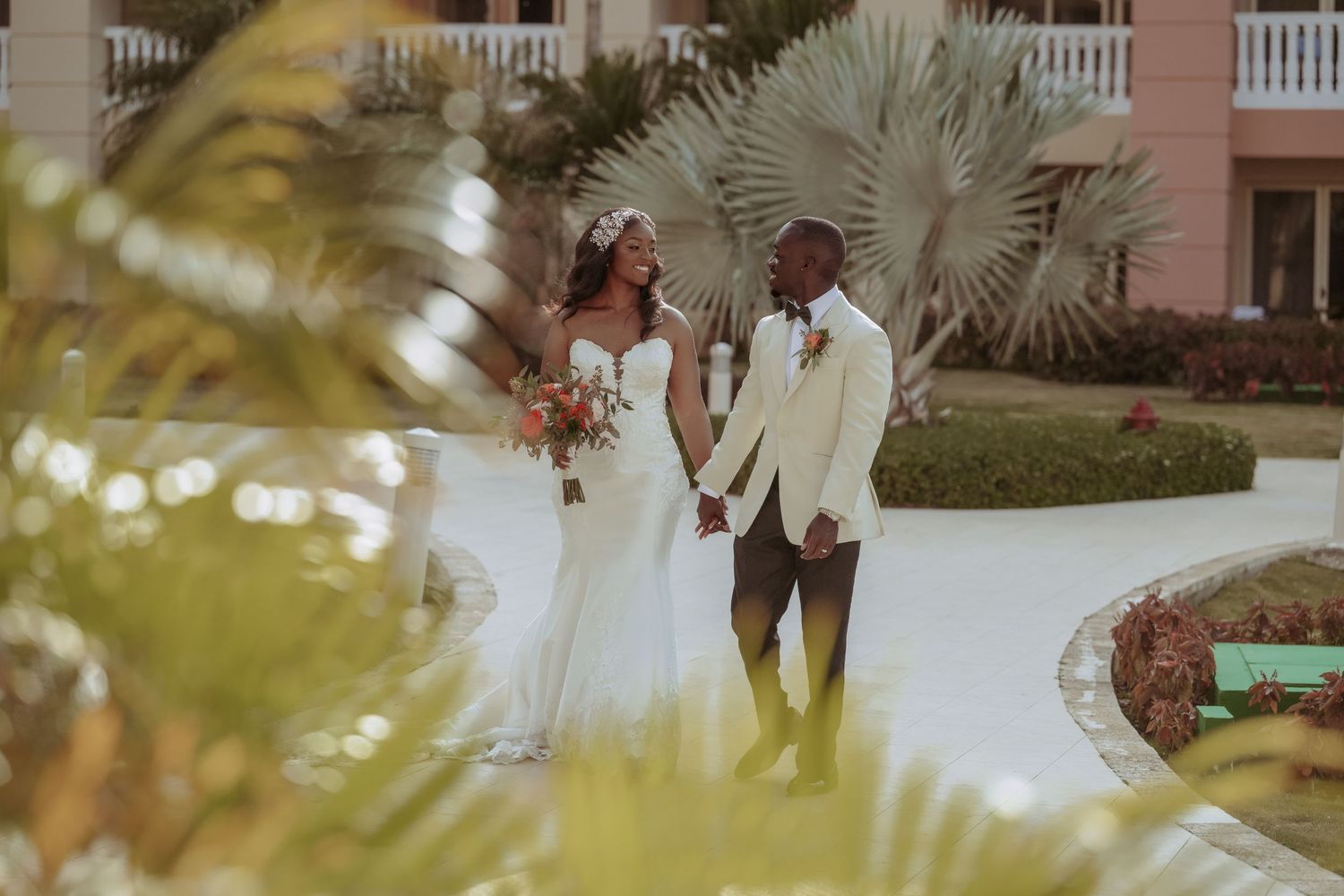 Romantic outdoor wedding ceremony at a tropical venue with palm trees and pink buildings in the background.