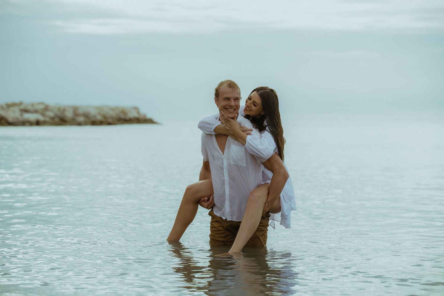 A couple plays and poses together in shallow tropical waters near a rocky coastline.