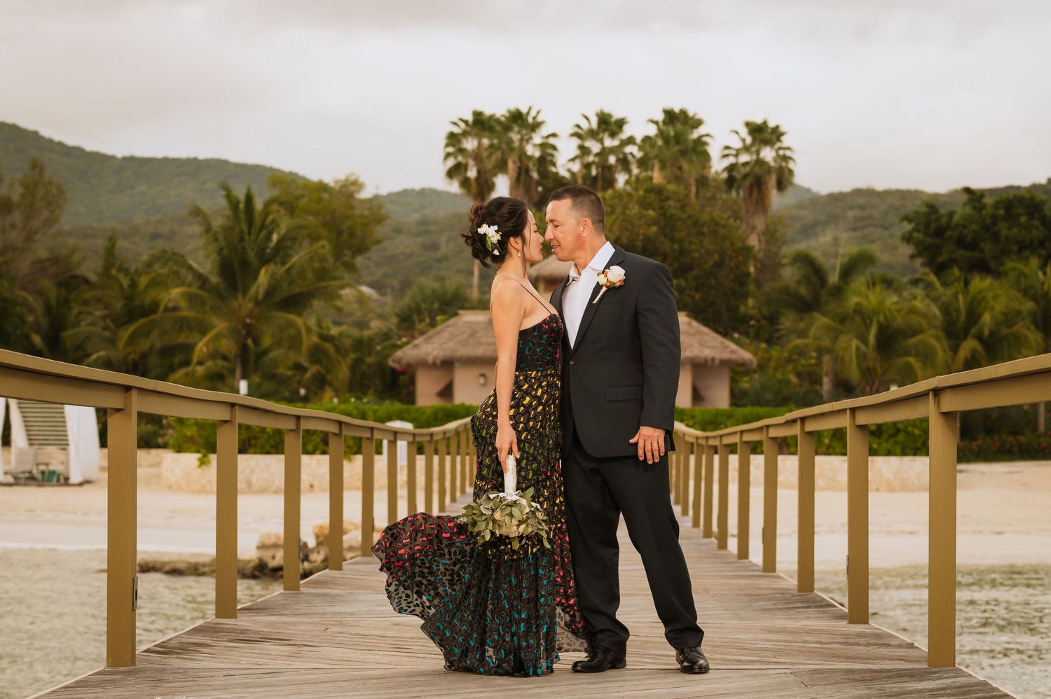 Couple poses on wooden boardwalk surrounded by palm trees and tropical landscape at dusk.