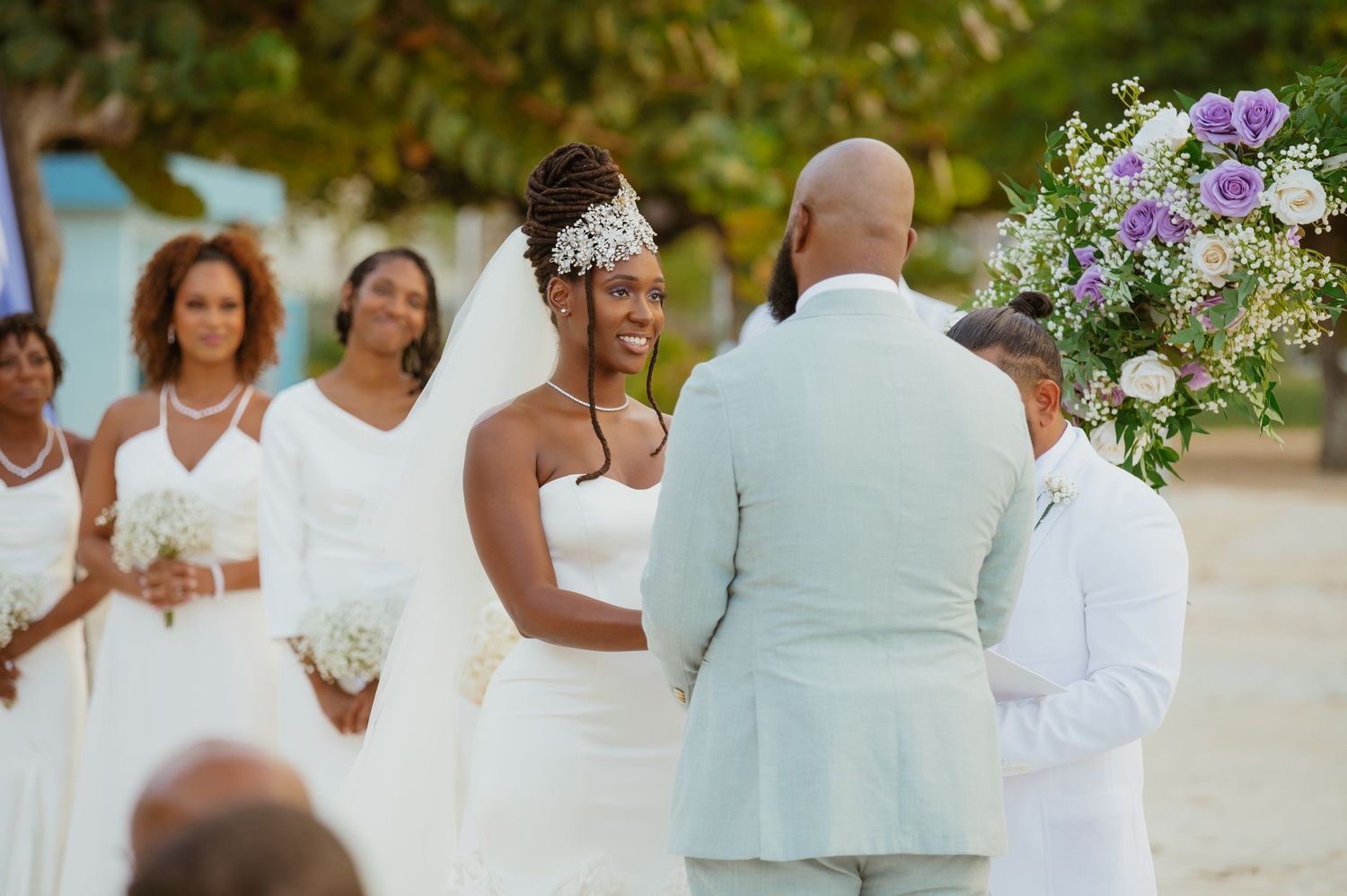 Wedding guests in white gather around a floral-decorated beach ceremony space.