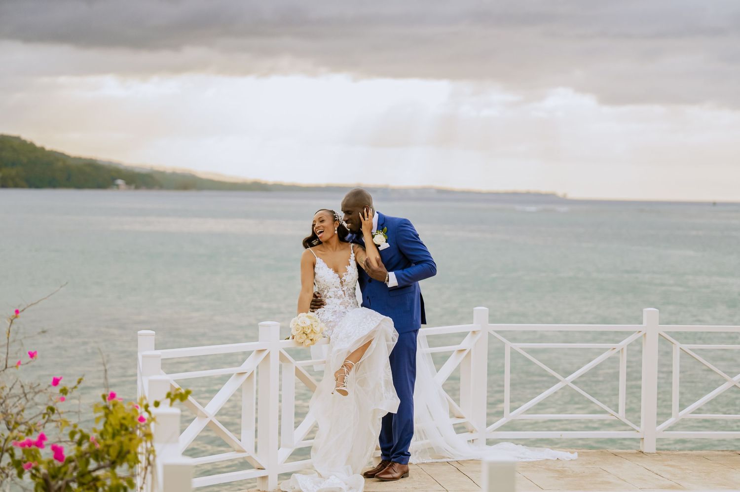 Coastal wedding scene with white deck railing, ocean views, and tropical flowers.