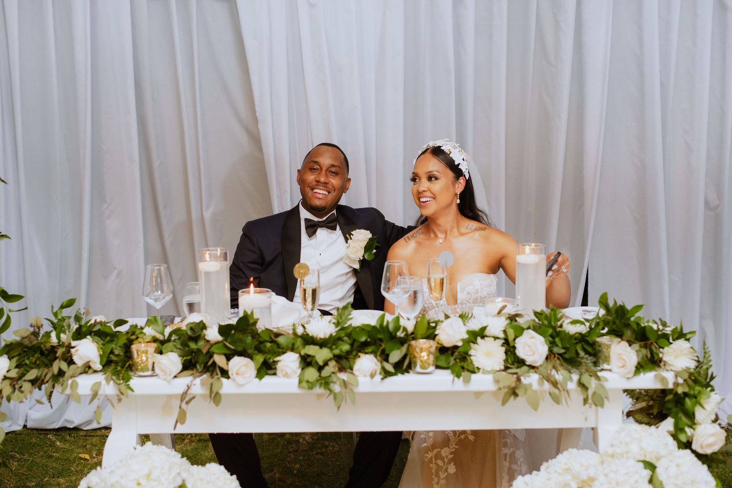 Newlyweds seated at elegant head table decorated with white floral arrangements and greenery.