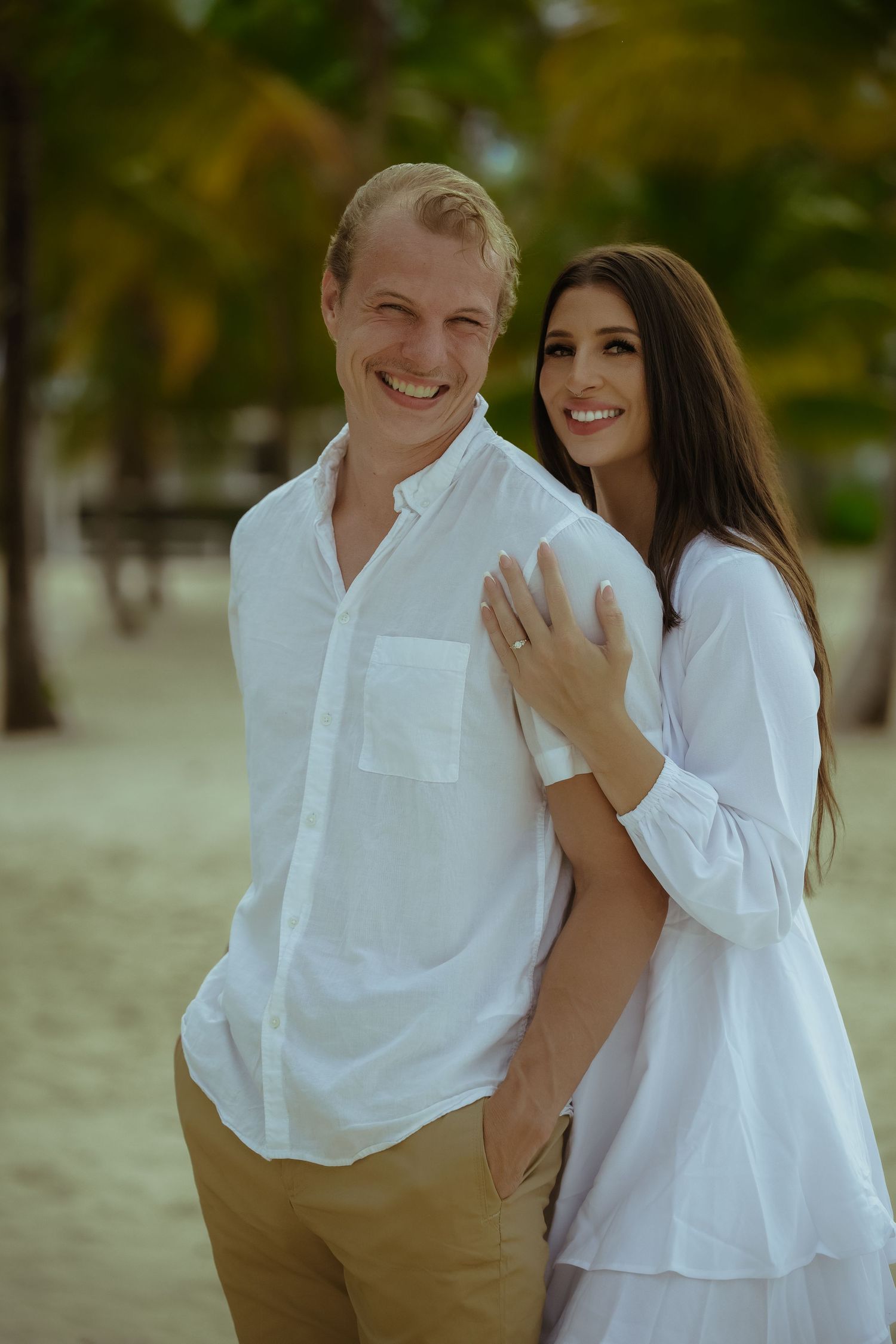 Couple dressed in coordinating white outfits embrace lovingly on a sandy beach with palm trees.