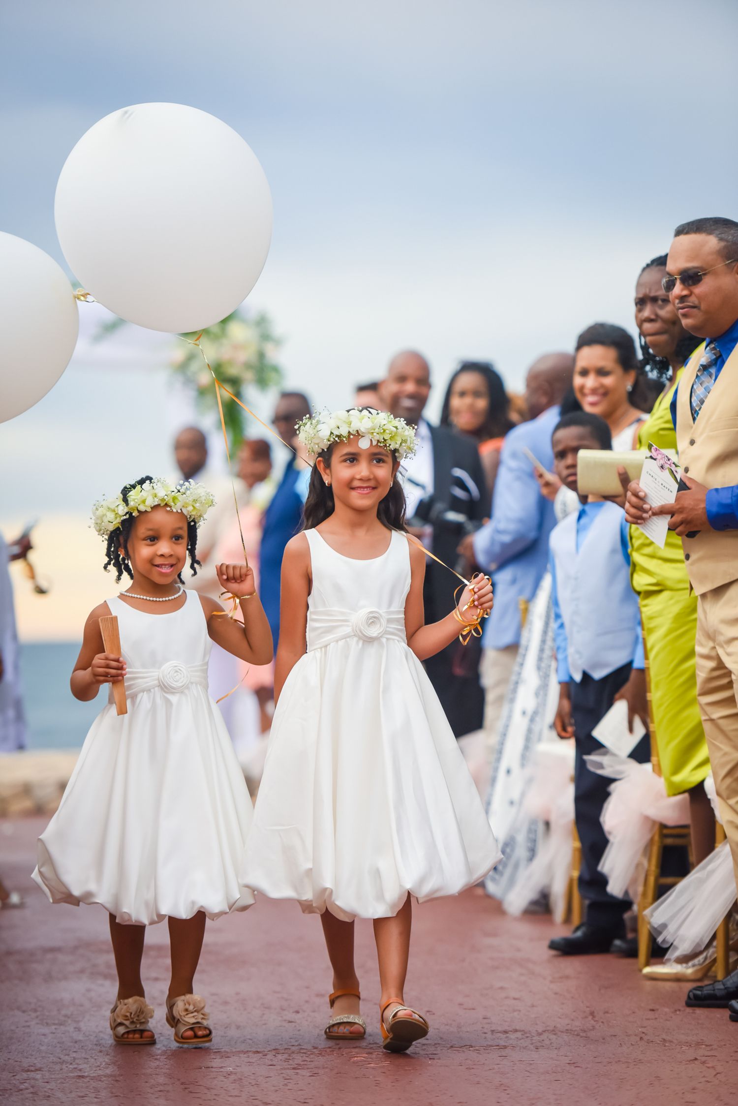 Two flower girls in white dresses and floral crowns walk down the aisle holding white balloons.