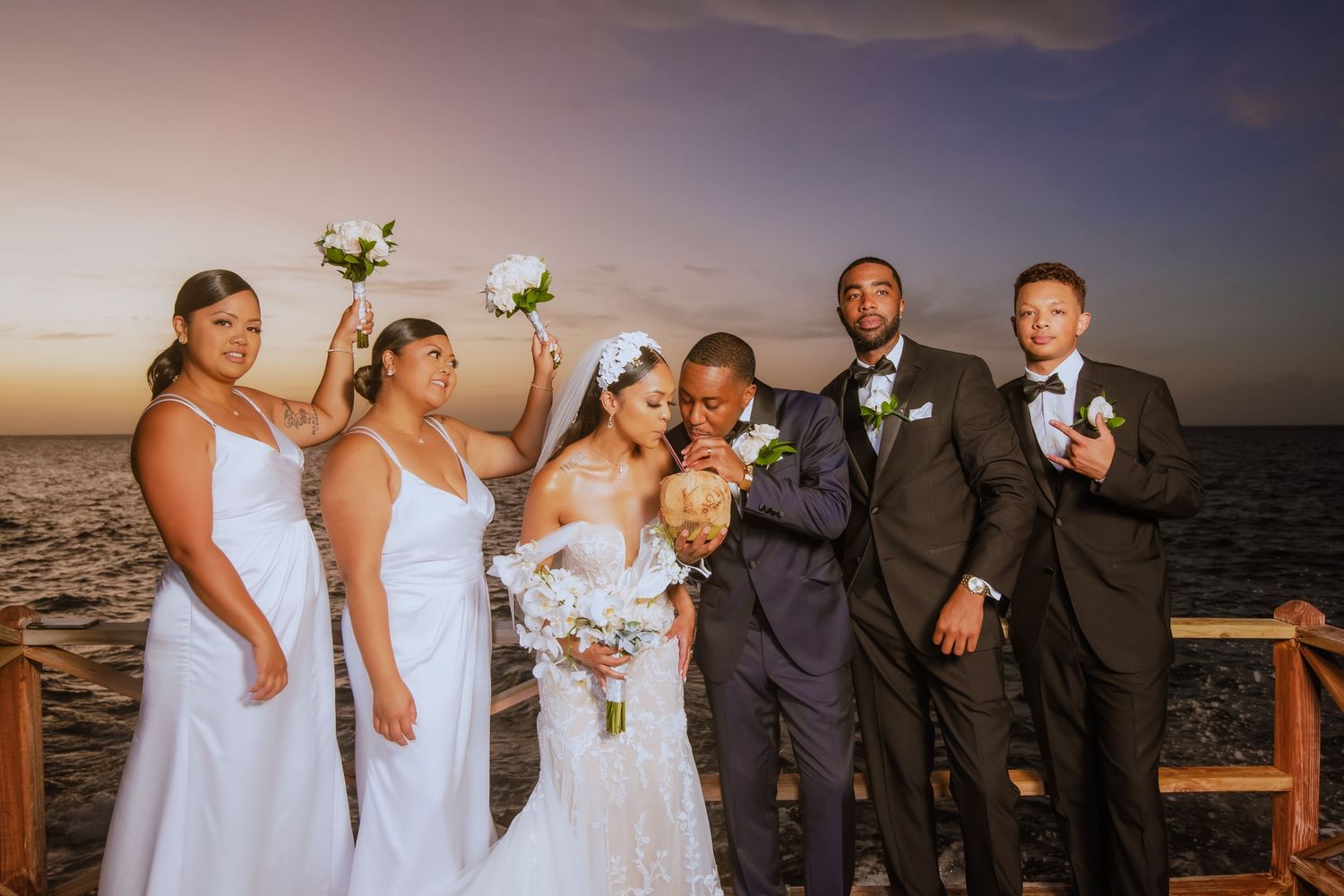 Wedding party poses together at sunset on oceanfront deck with white flowers and formal attire.