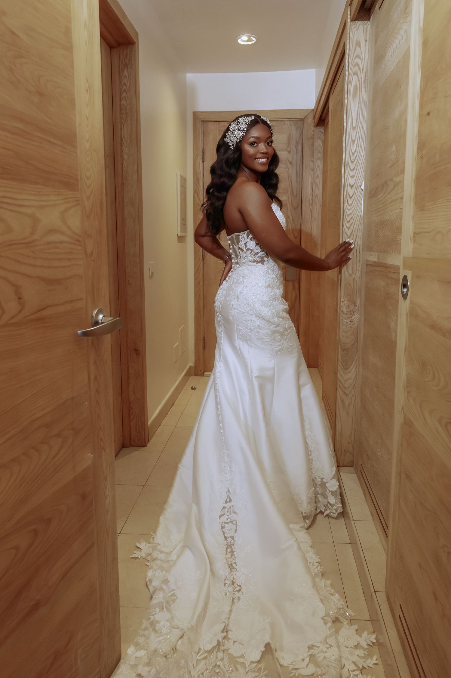 Elegant bridal portrait showcasing detailed lace train and crystal headpiece in wooden hallway.