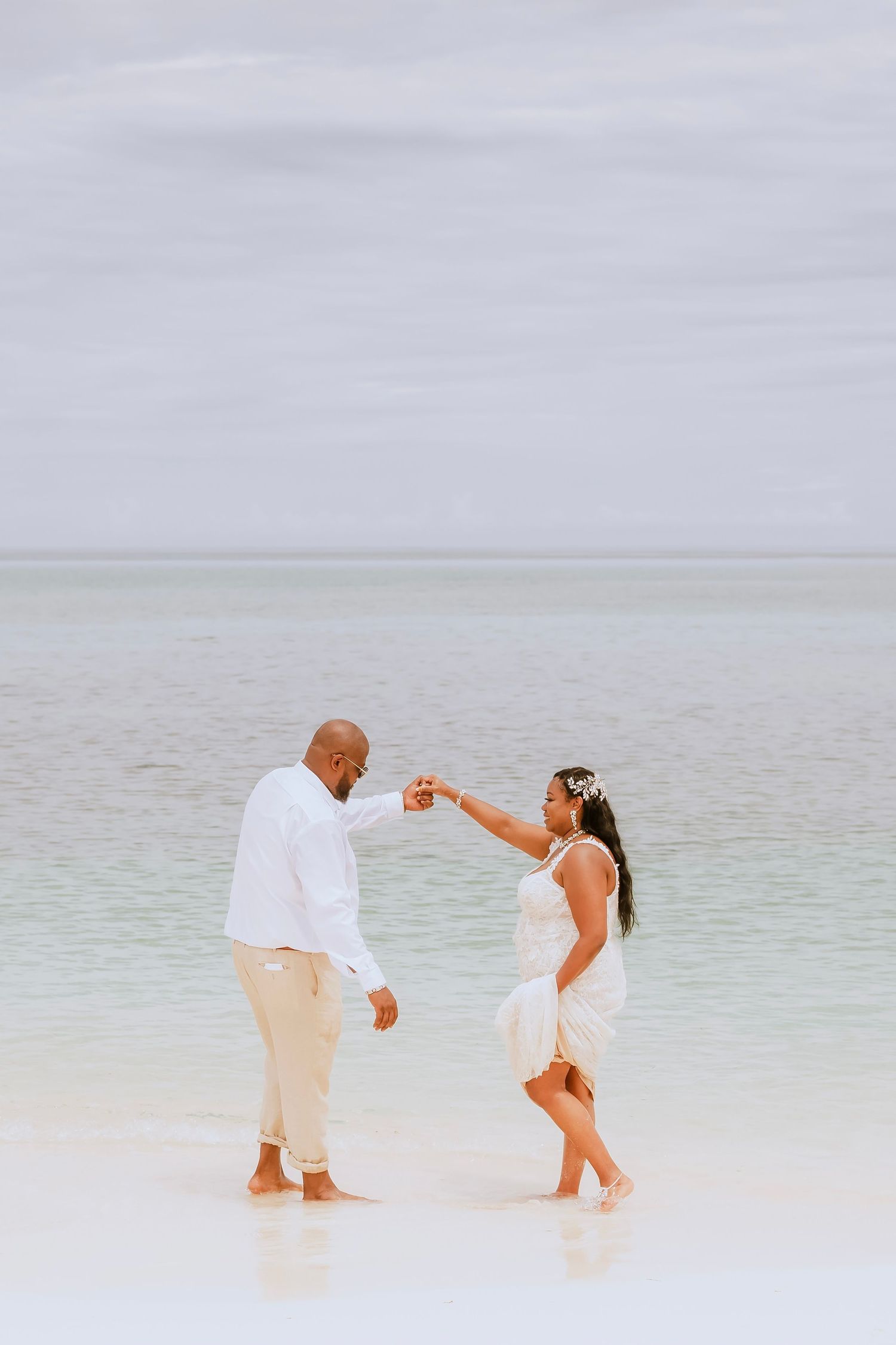 Couple in white attire dances playfully along pristine beach shoreline.