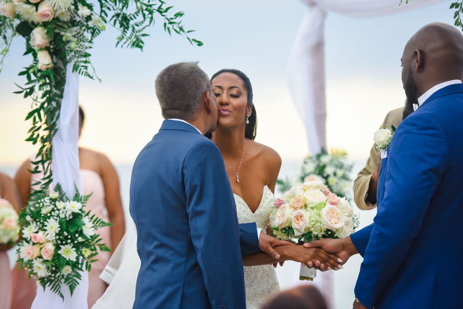 A tender moment unfolds during a wedding ceremony beneath an ornate floral arch with greenery.