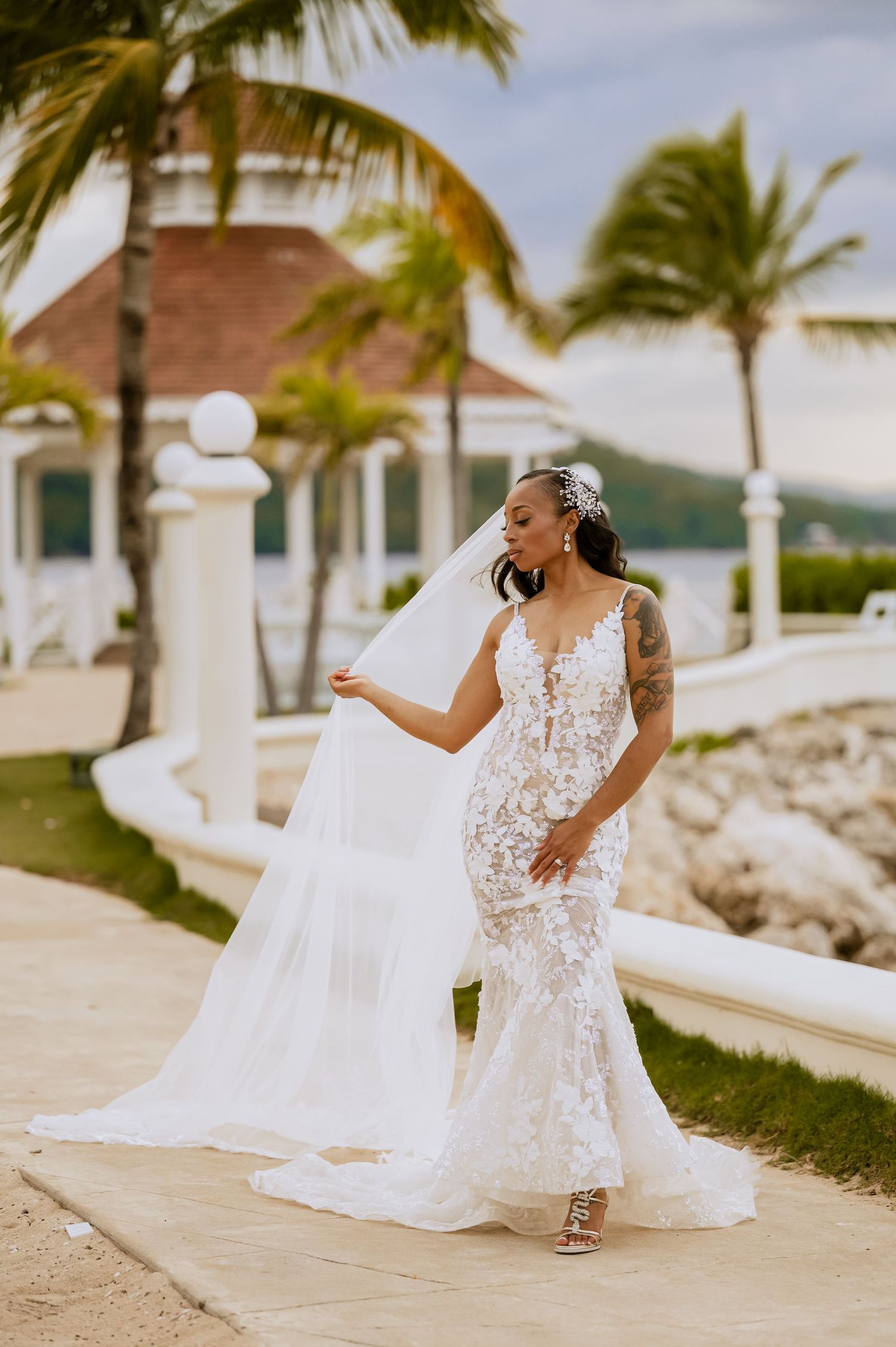 A bride in an elegant lace gown dances on a seaside walkway surrounded by palm trees.