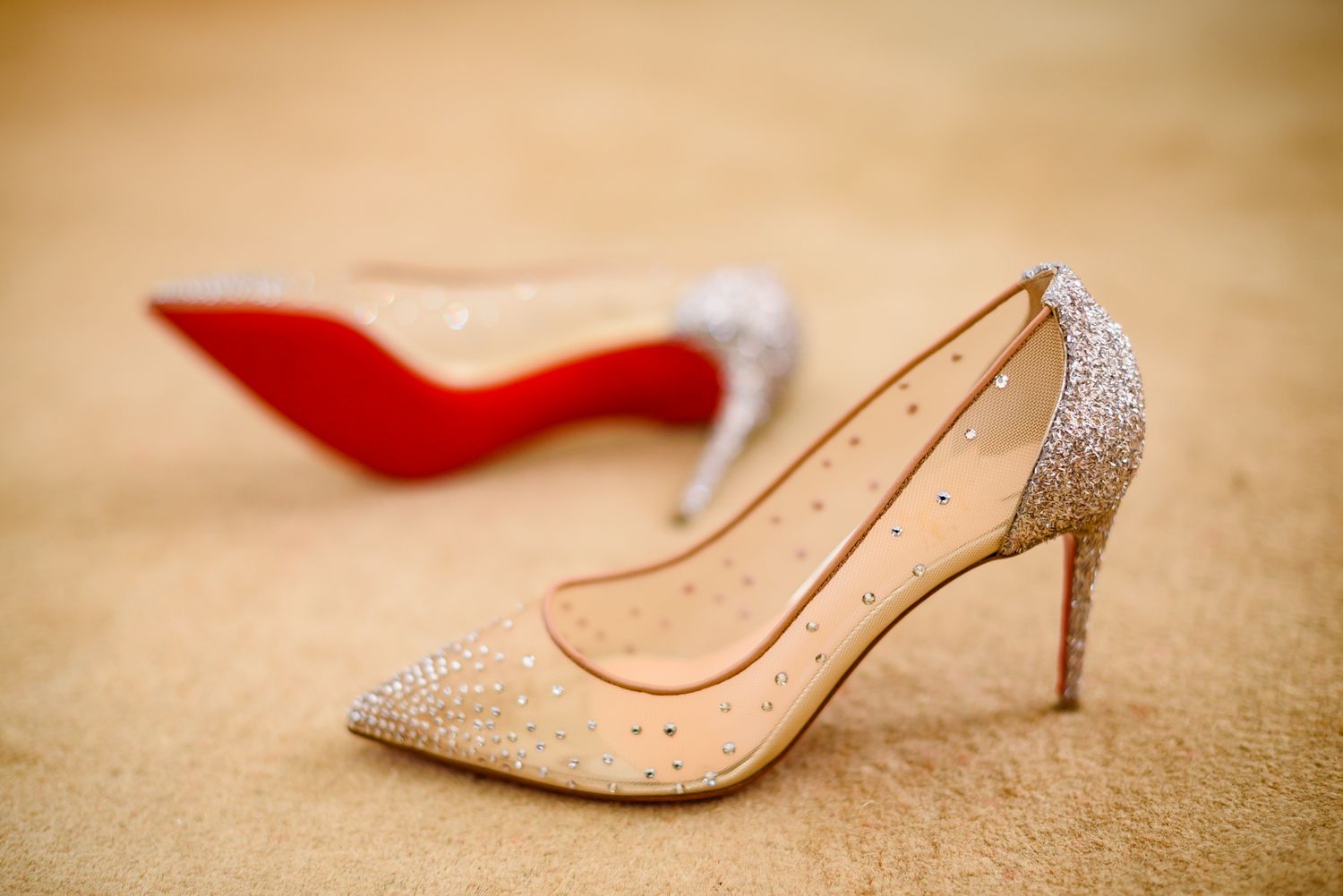 A sparkly silver and nude stiletto heel sits next to a red-soled shoe on a beige carpet.