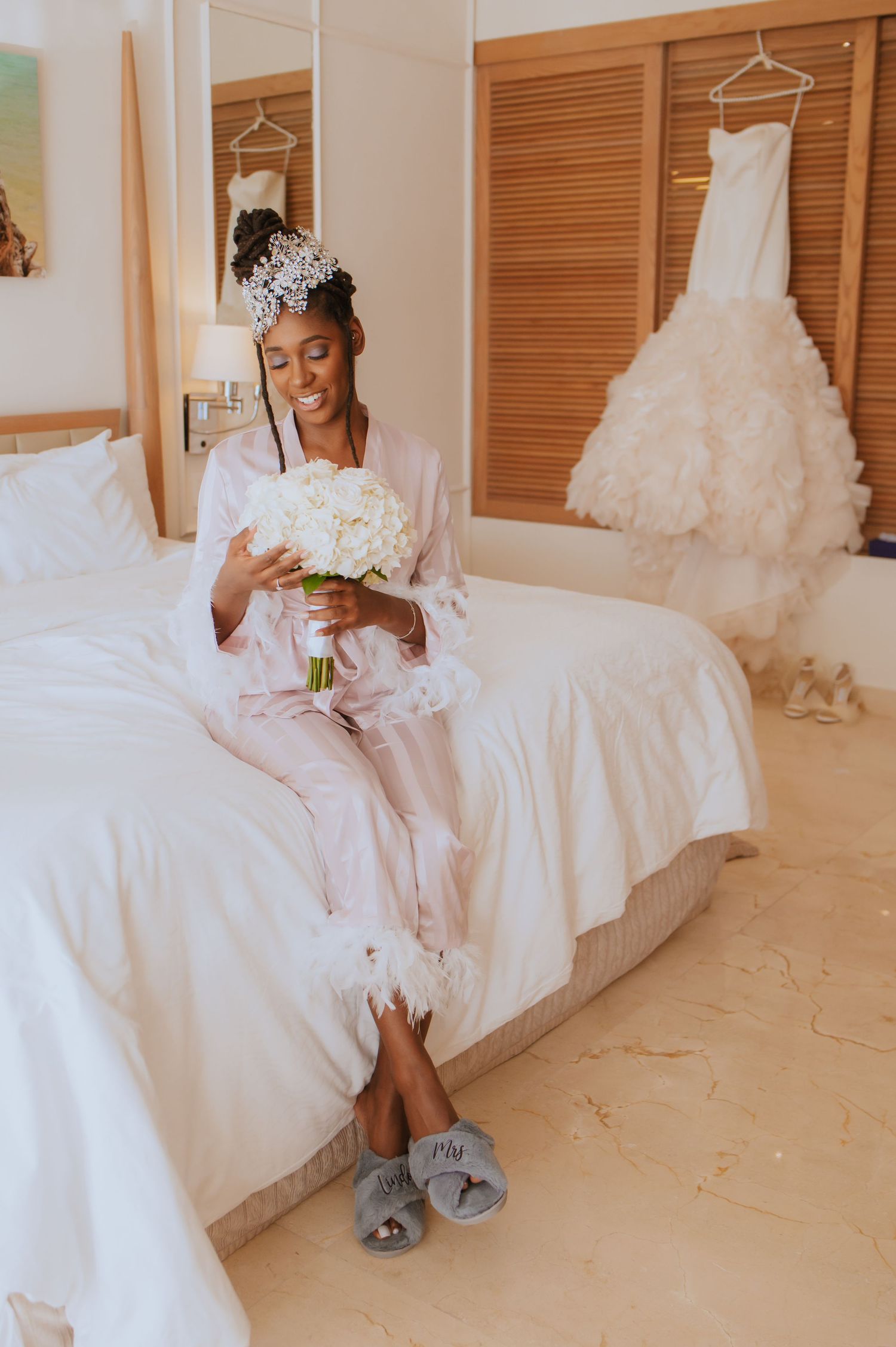 Bride in robe sits on bed with white bouquet and wedding dress visible in background.