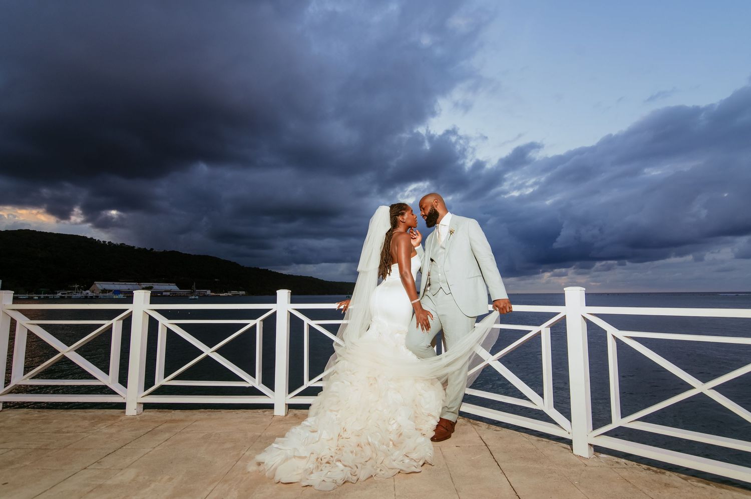 A bride in a ruffled wedding gown leans gracefully against a white balcony railing with dramatic clouds behind.