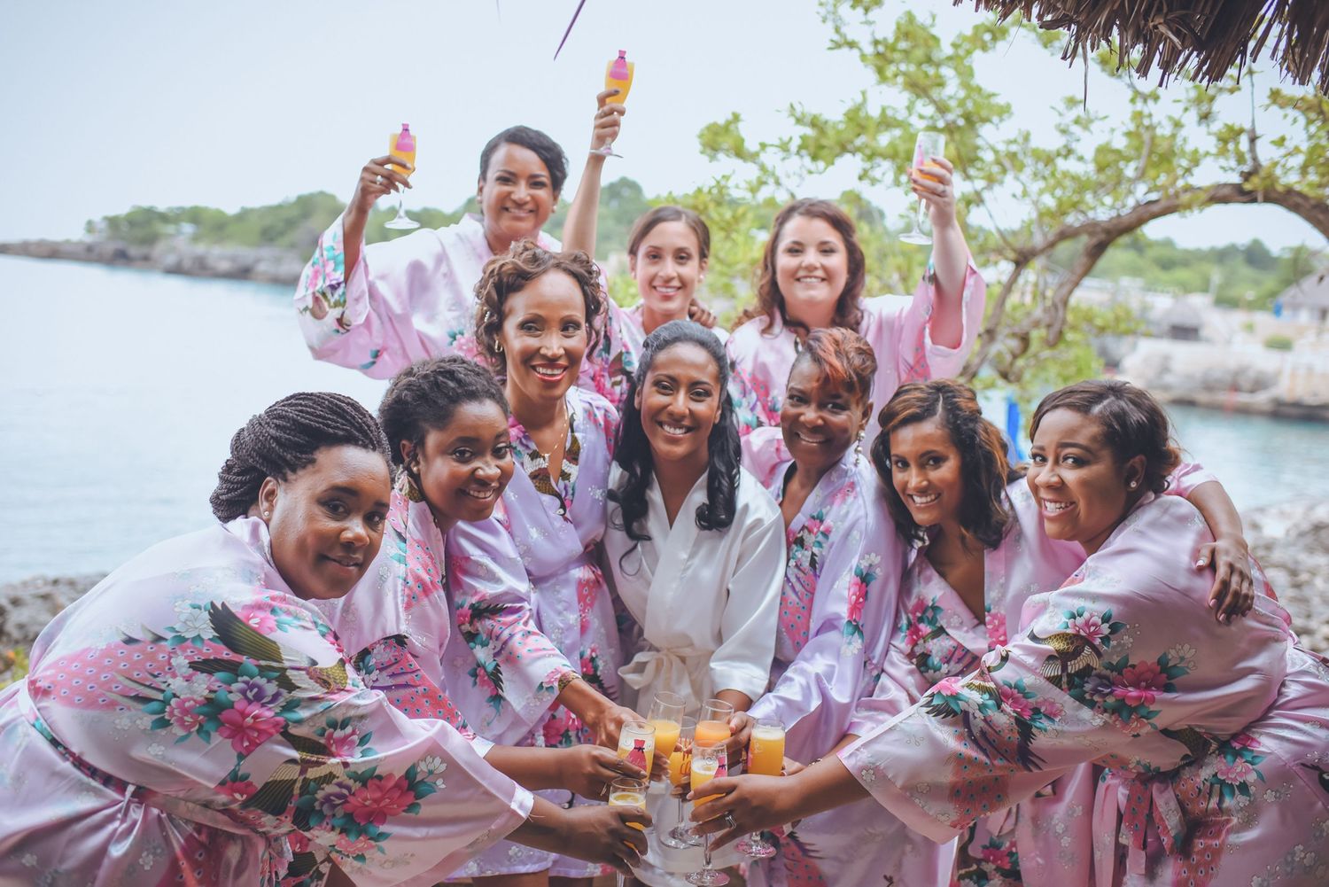 Group wearing floral robes celebrates on tropical beach during pre-wedding festivities.