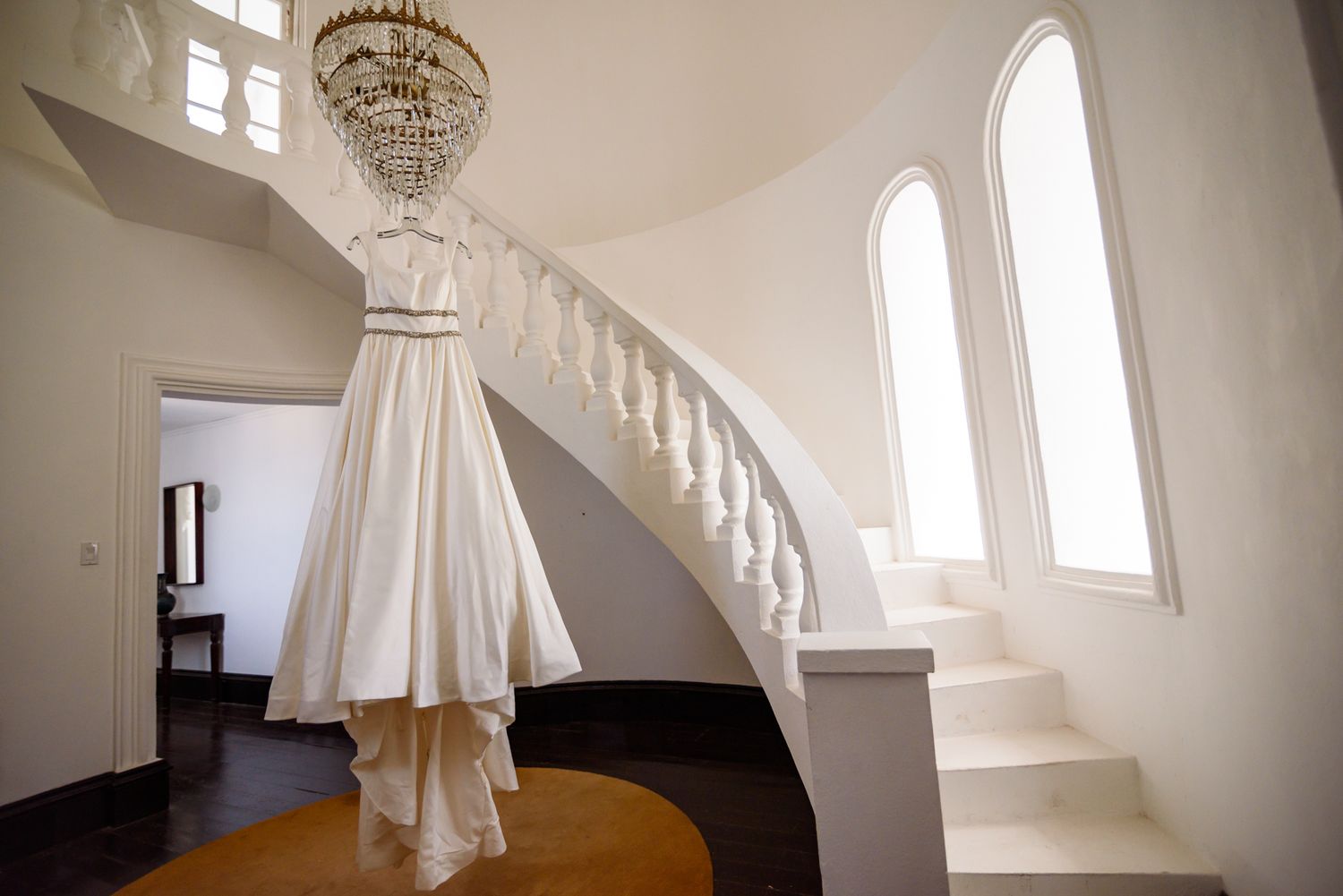 A white wedding dress hangs from a crystal chandelier near a curved white staircase with arched windows.