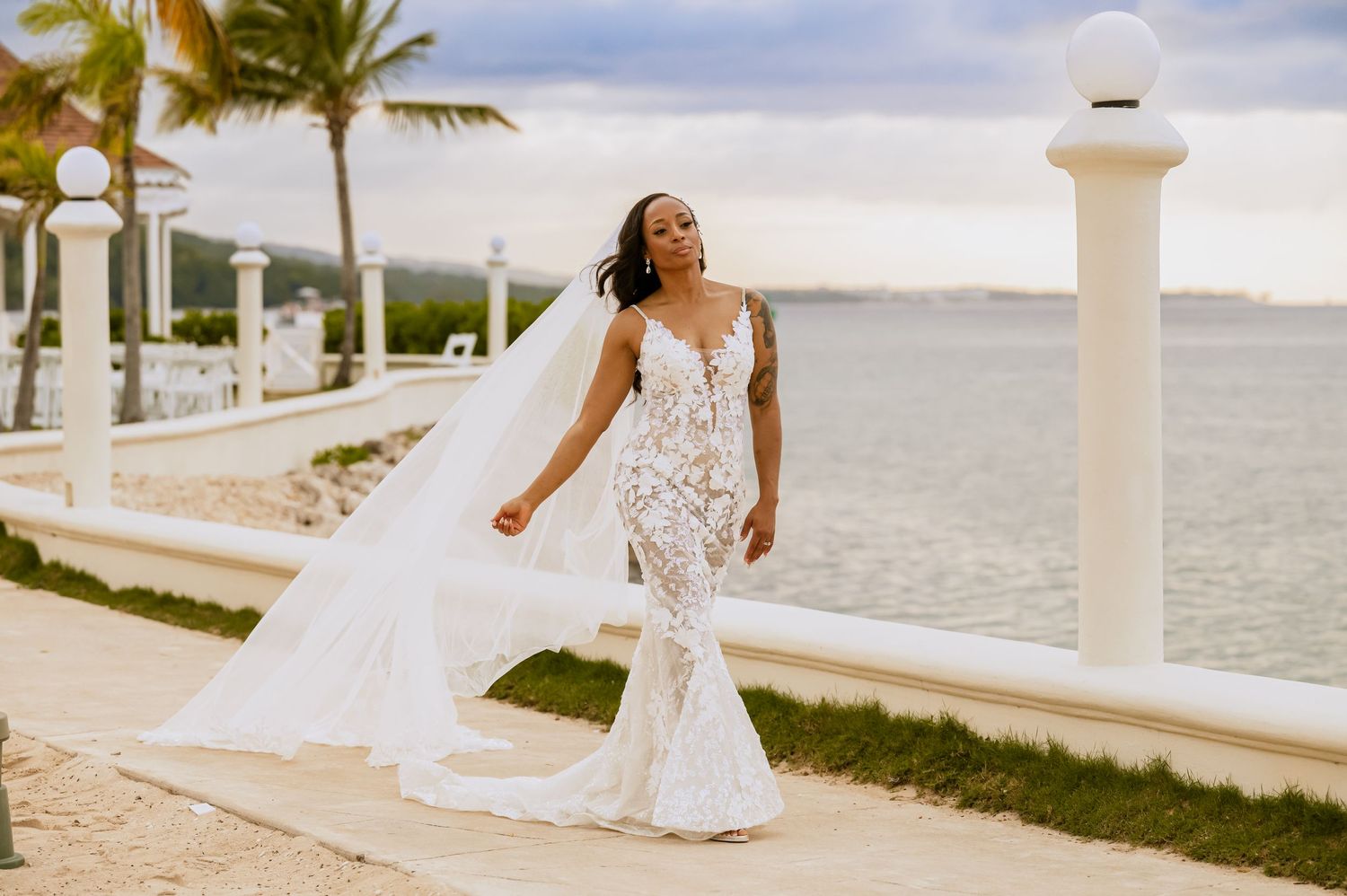 A wedding dress with a flowing veil photographed against a waterfront backdrop with palm trees and white pillars.