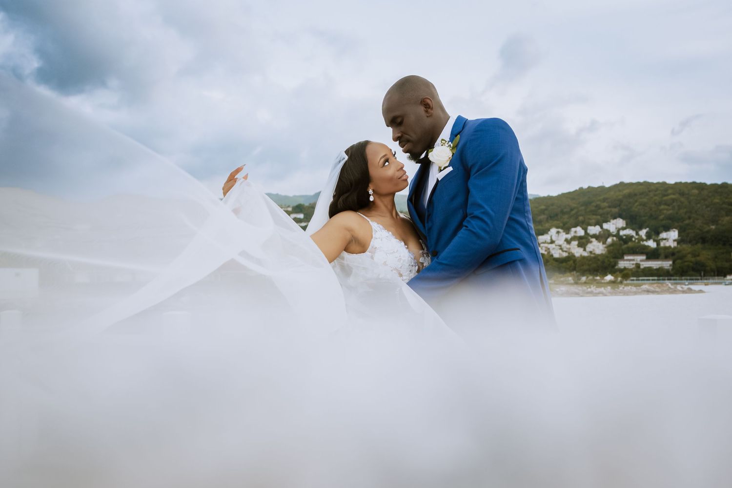Romantic wedding photo with flowing veil against cloudy sky and scenic landscape.