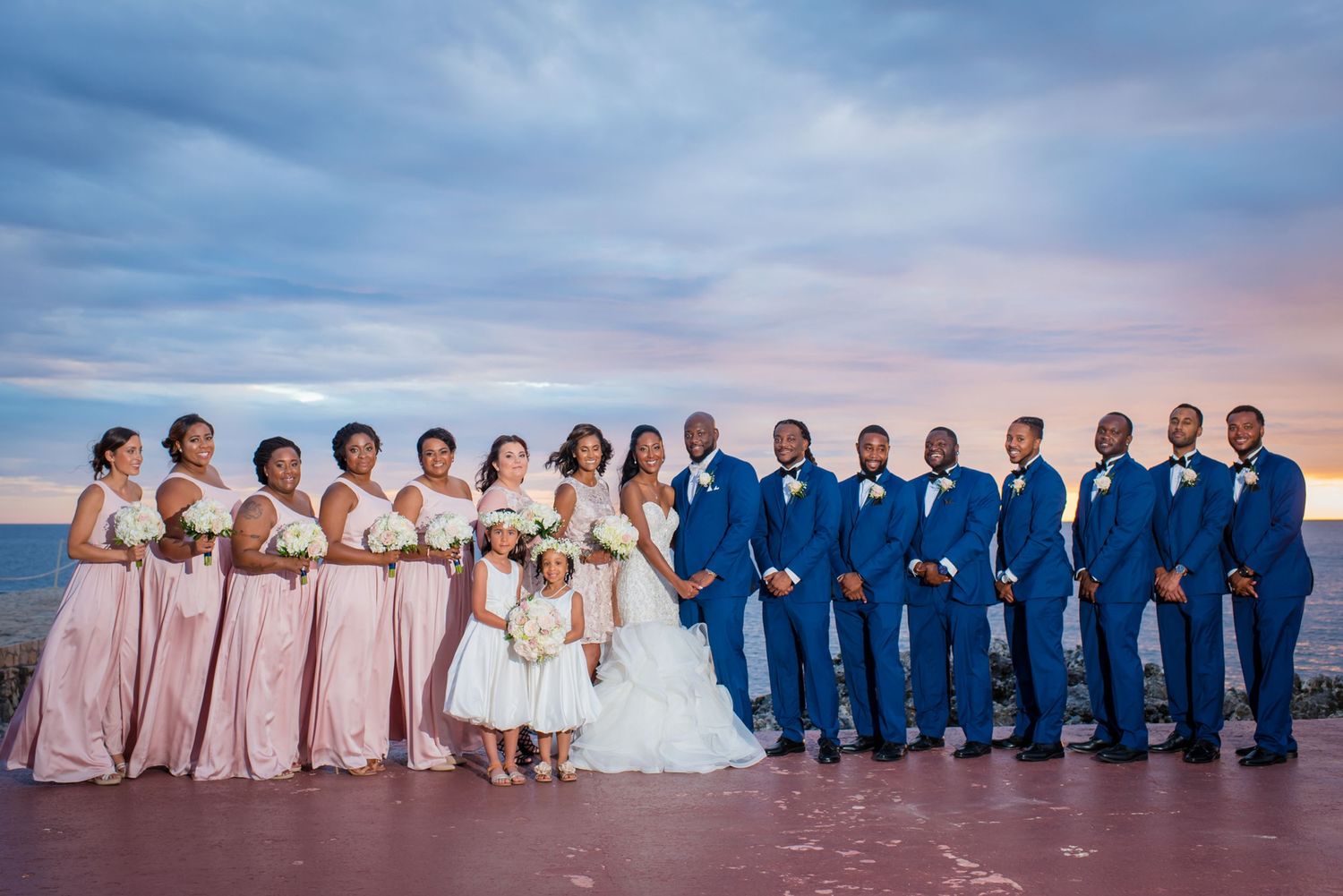 Wedding party in blue and pink attire poses at sunset with ocean backdrop.