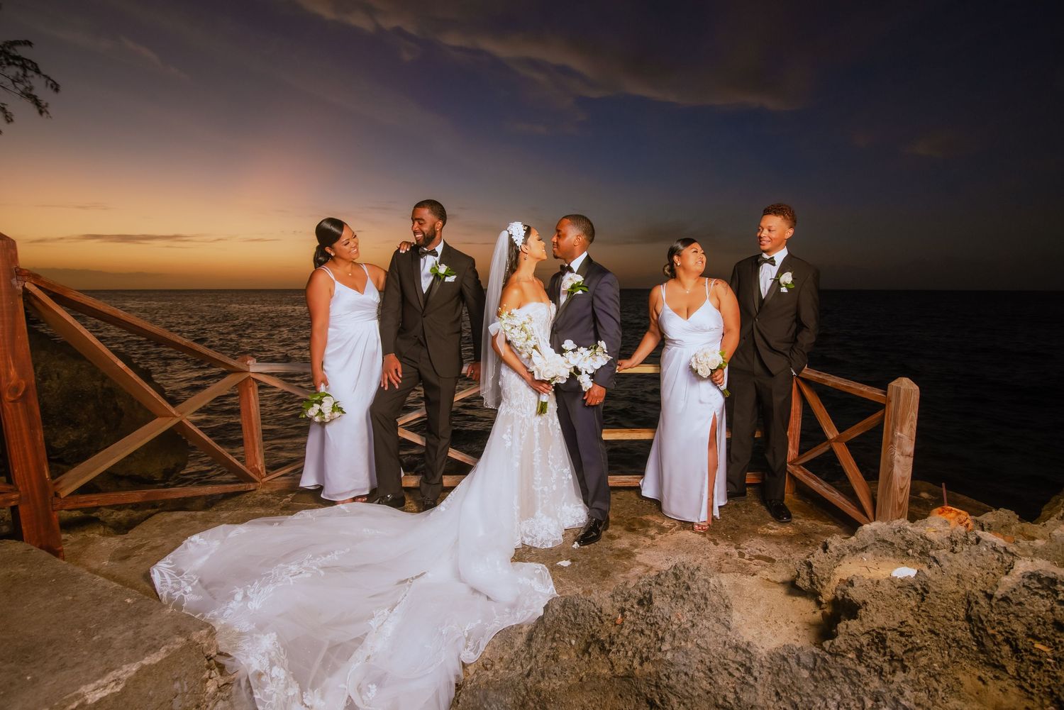 Elegant wedding party gathers on coastal overlook during golden hour.