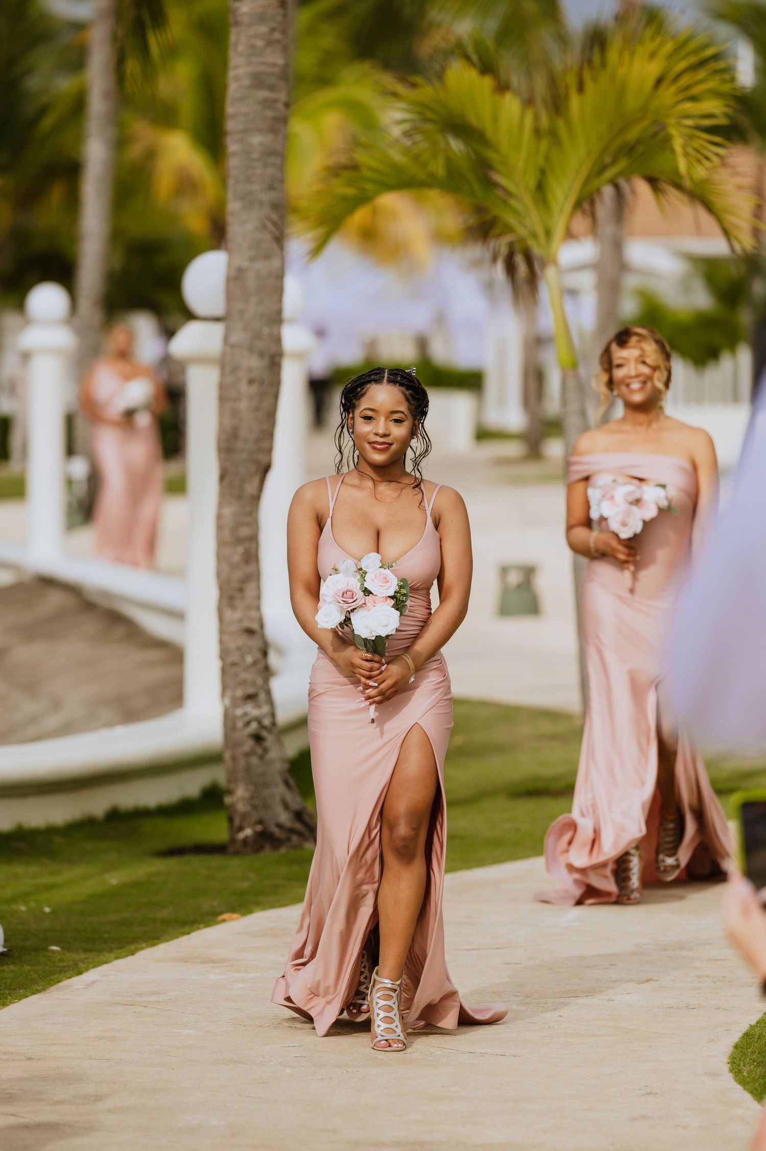 Bridesmaids in blush pink dresses hold bouquets while walking along tropical pathway.