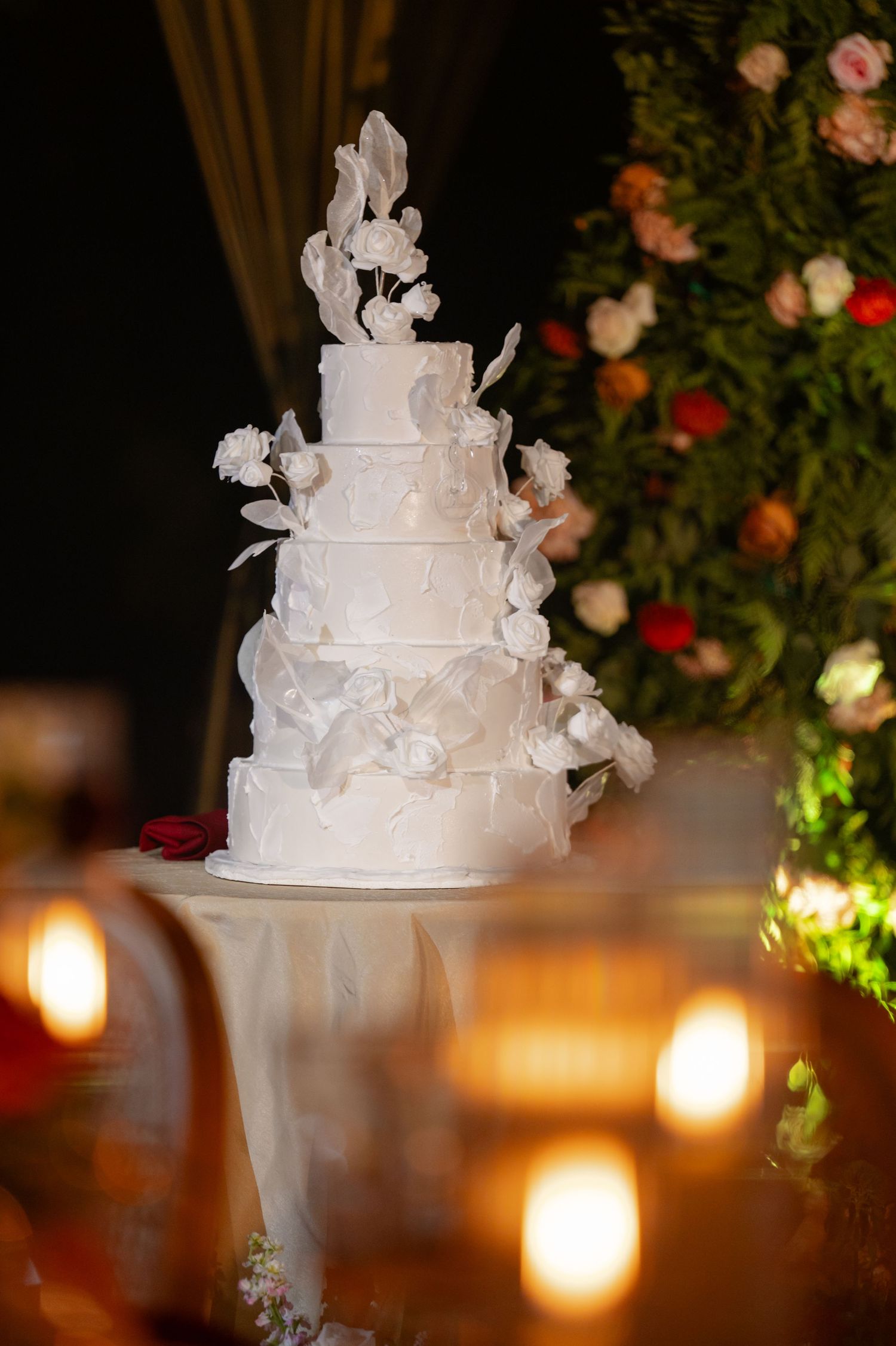 Elegant white wedding cake decorated with delicate sugar flowers and butterflies, illuminated by warm candlelight.