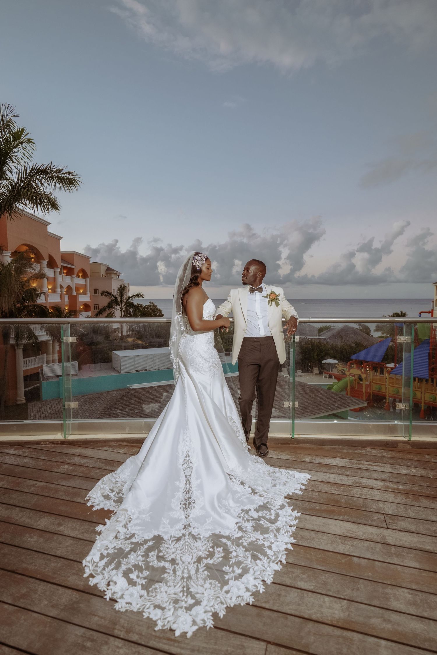 Wedding couple on wooden deck overlooking ocean with palm trees and resort buildings in background.