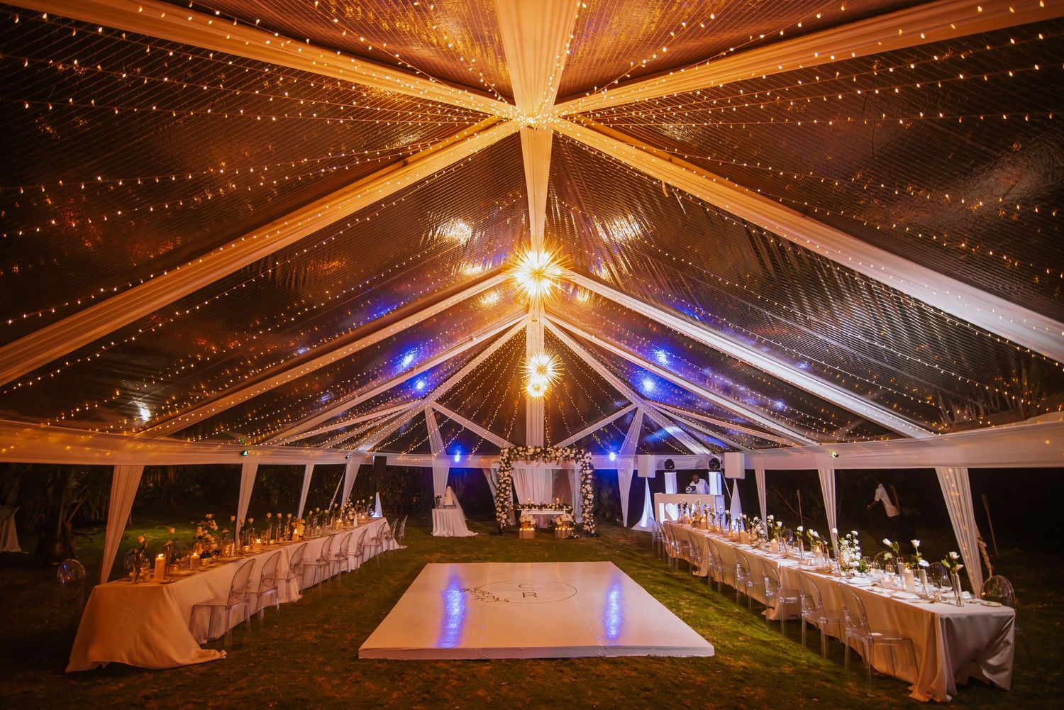 Clear tent wedding reception venue with string lights creating a starry ceiling effect and white table settings.