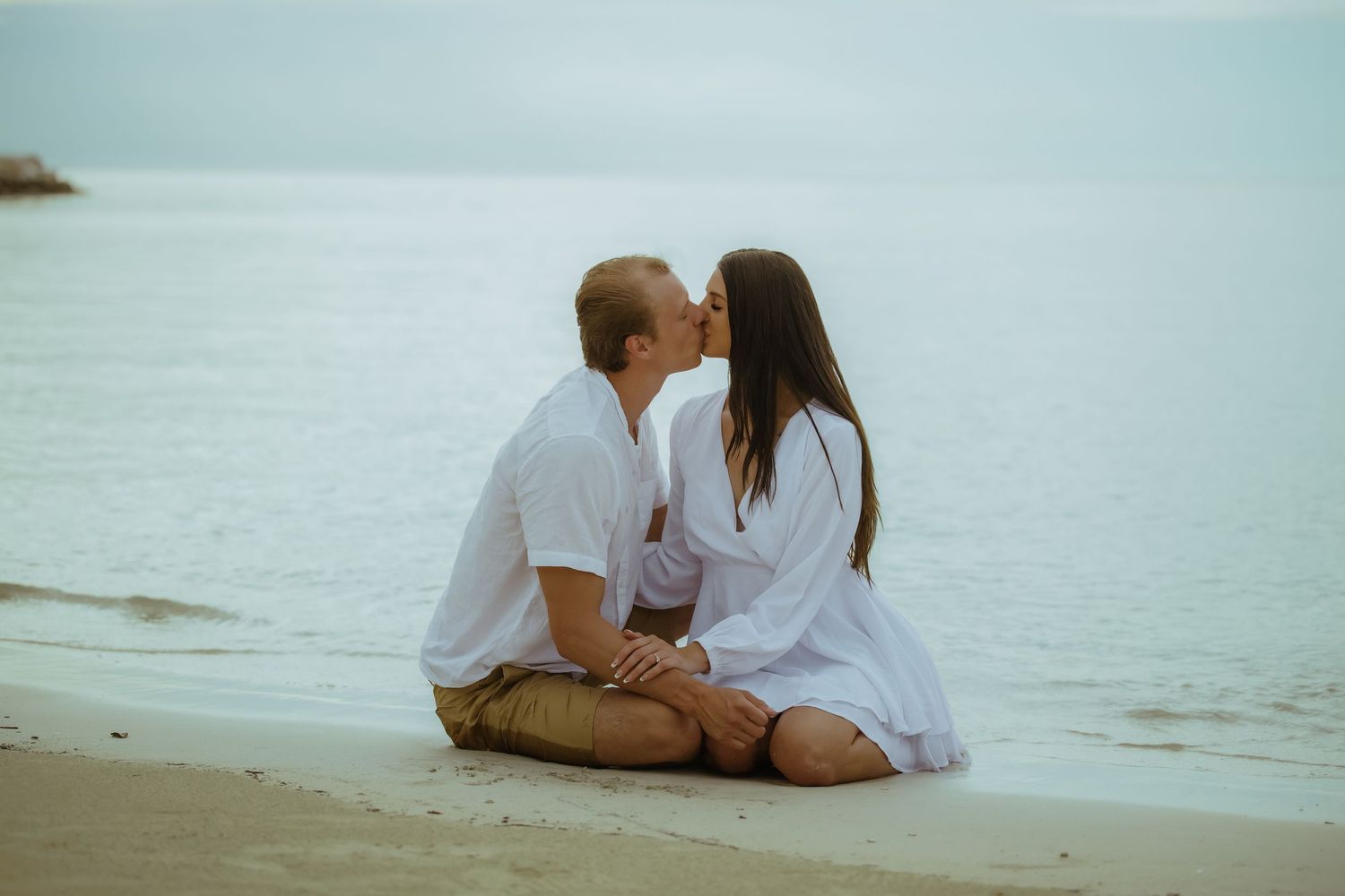 A couple in matching white outfits shares an intimate moment while sitting on a beach.