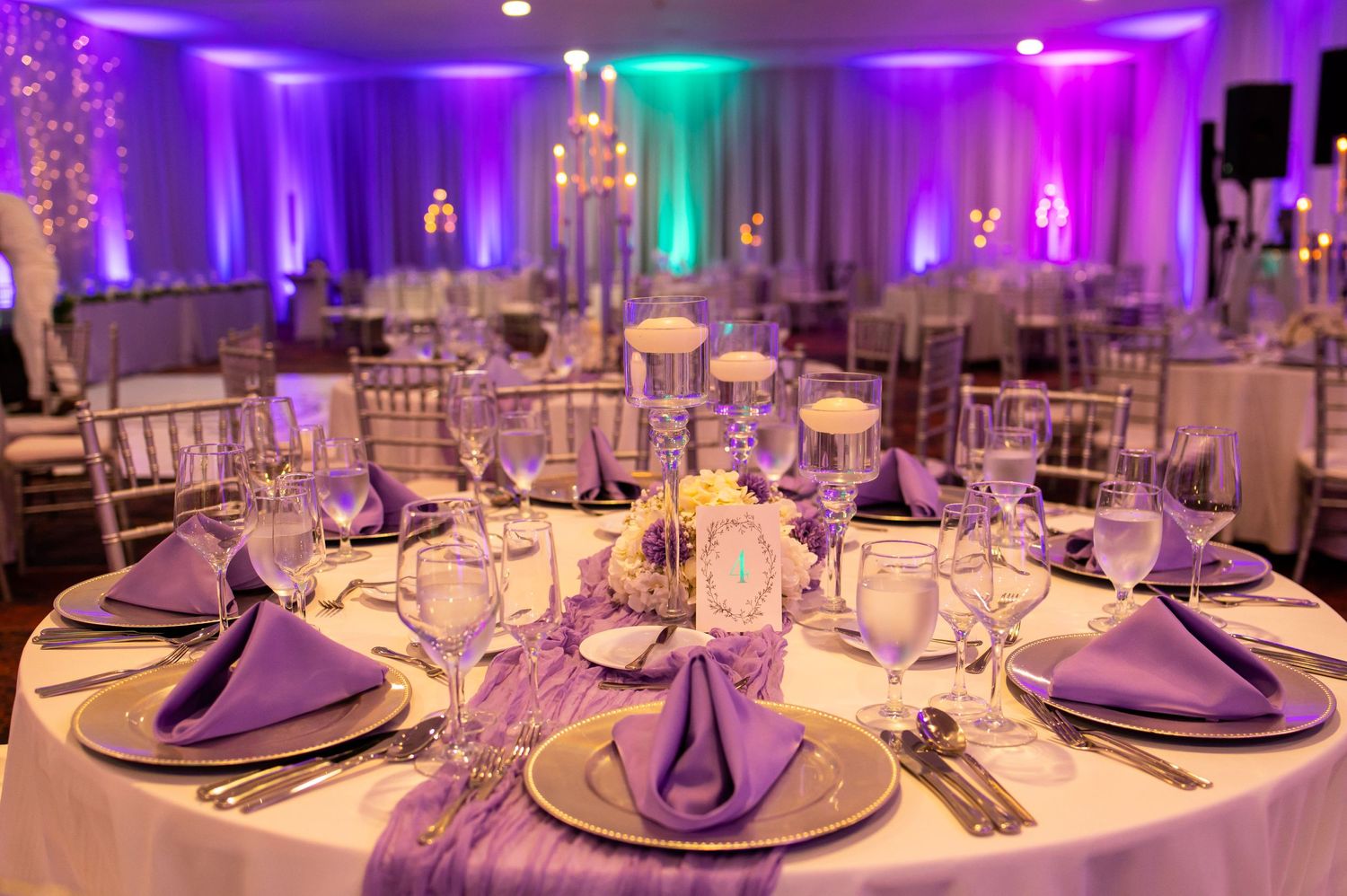 Wedding reception tables with purple napkins and elegant place settings under mood lighting.