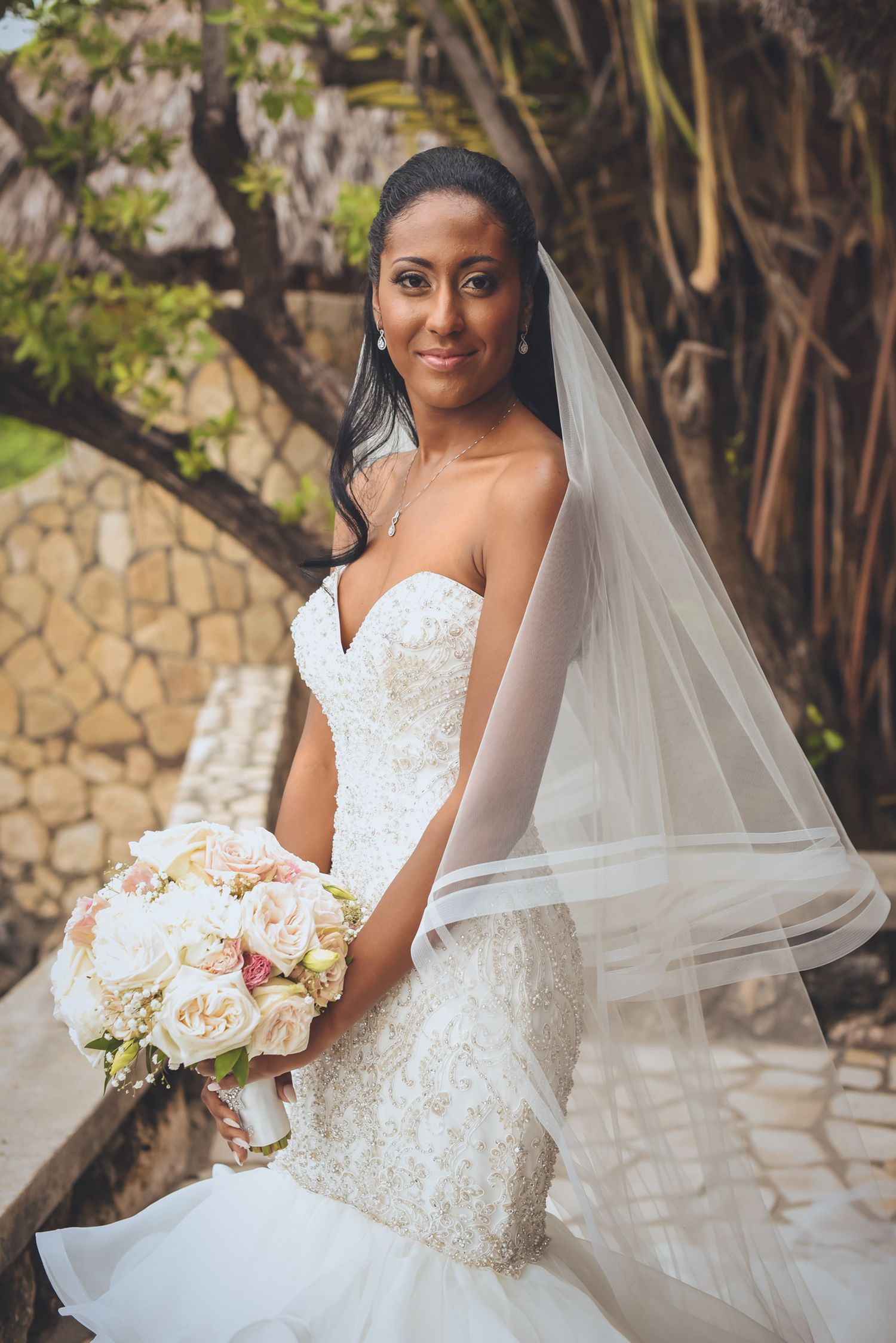 Elegant bride in a strapless white gown holds a cream and blush bouquet in a garden setting.