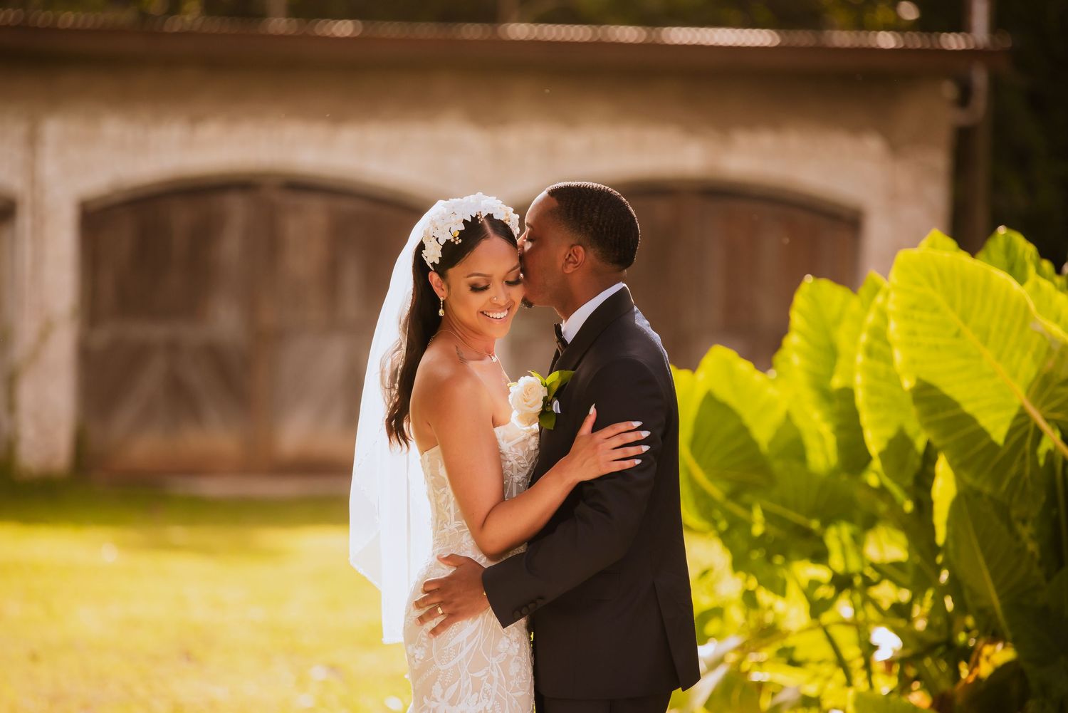 A romantic wedding couple embraces in golden sunlight with lush greenery in the background.
