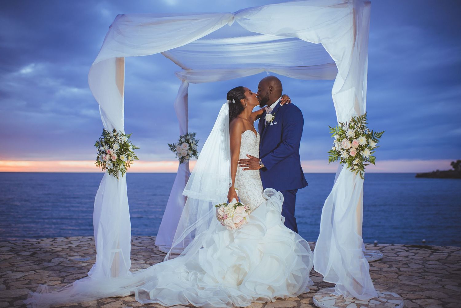 Oceanfront wedding ceremony setup with white fabric pavilion and tropical flower arrangements.