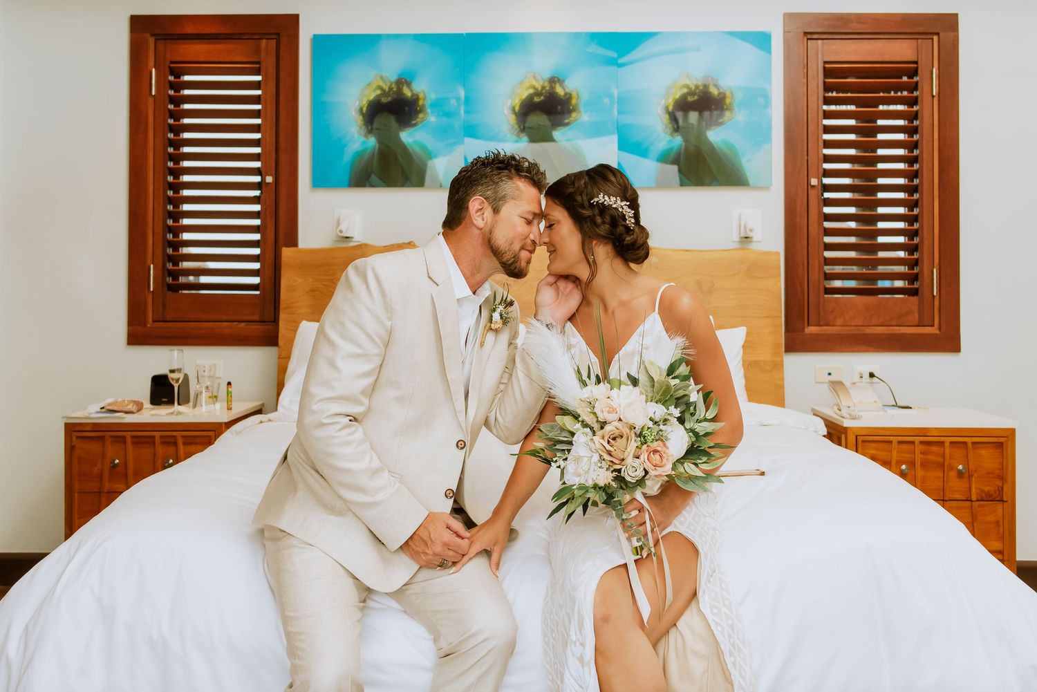 A romantic wedding scene on a white bed with wooden shutters and tropical decor in the background.