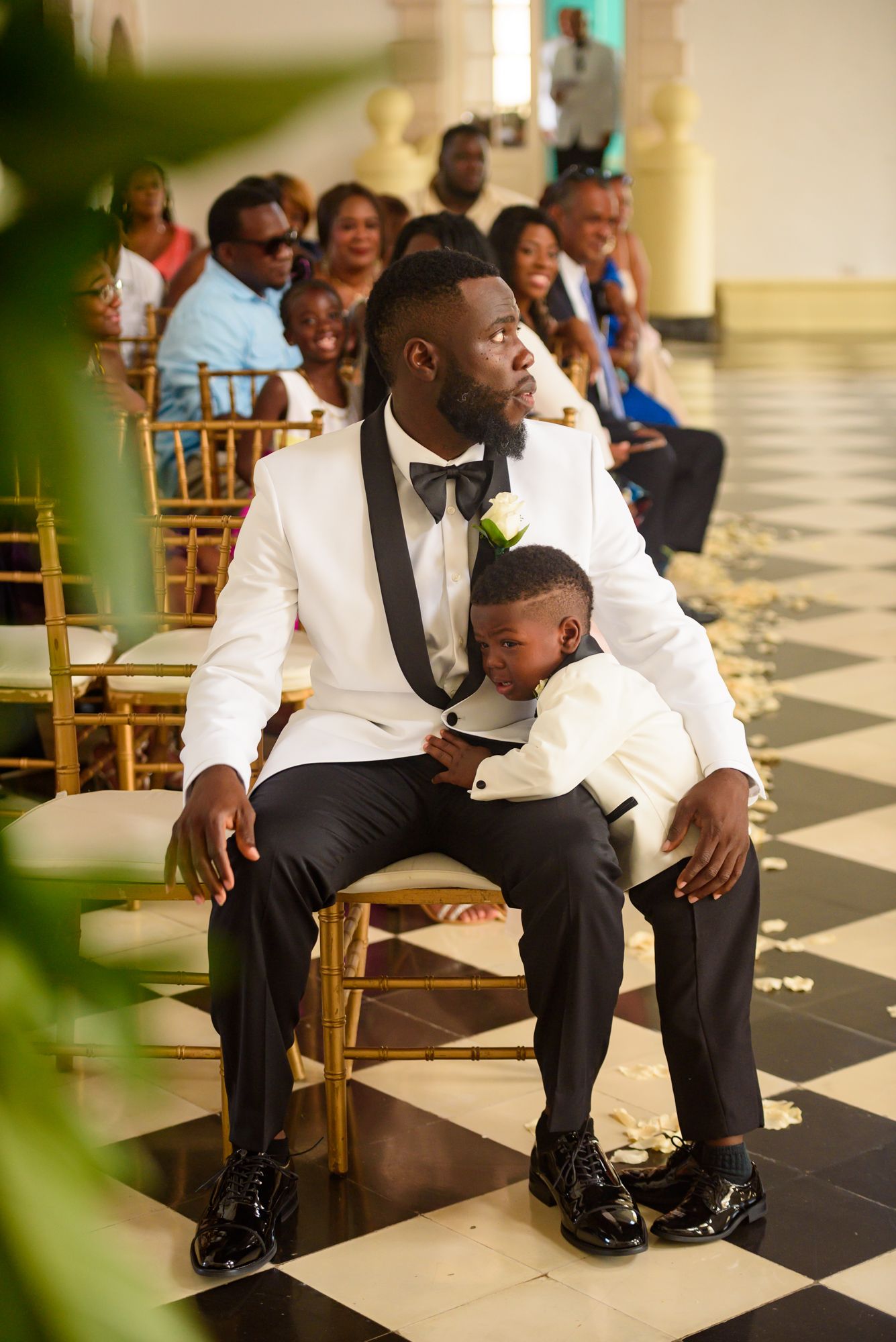 Wedding party members in white tuxedos seated on gold chairs in an elegant venue.