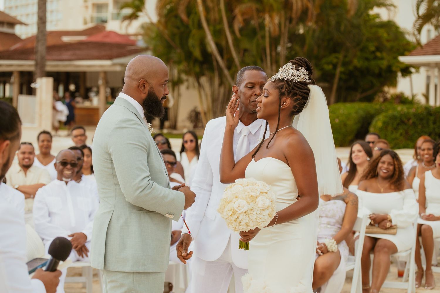 Joyful wedding celebration on a palm-lined beach with guests in coordinated white outfits.