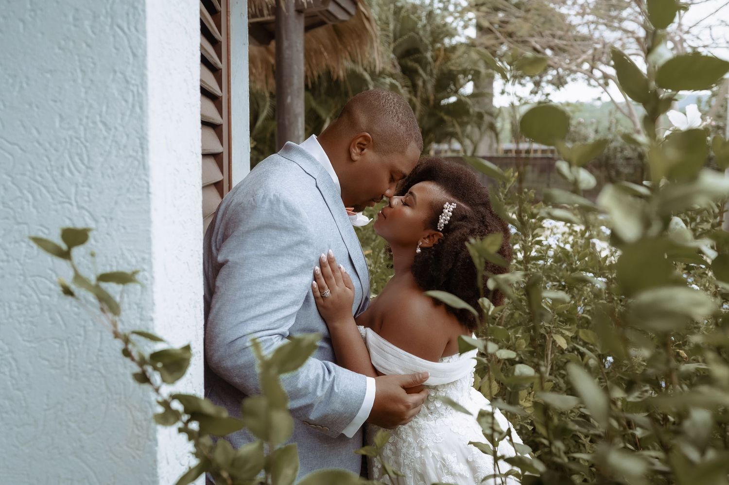 A couple shares an intimate moment in a garden setting surrounded by lush greenery and white walls.