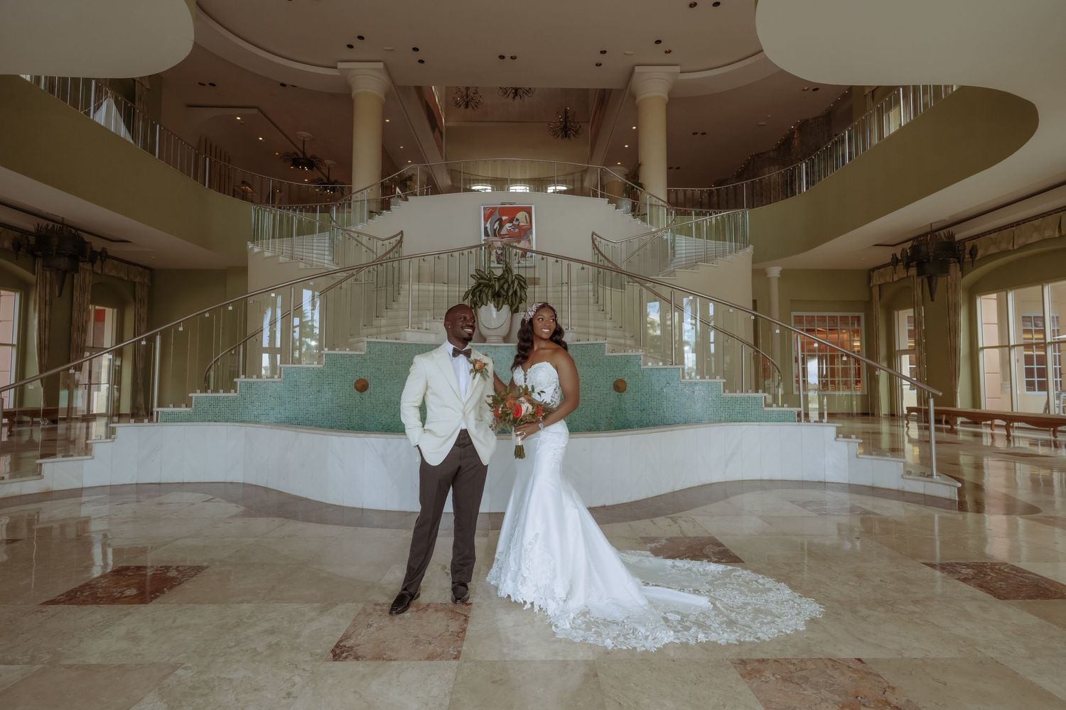 A couple in wedding attire poses in front of a grand staircase and fountain in an elegant indoor venue.