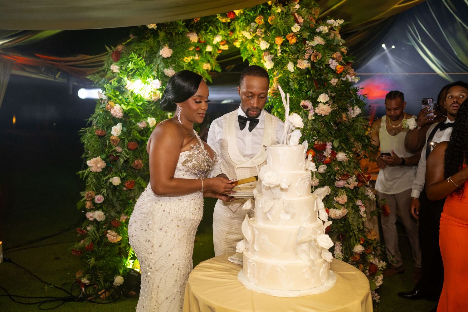 Elegant white wedding cake display with floral decorations and greenery at an outdoor evening reception.