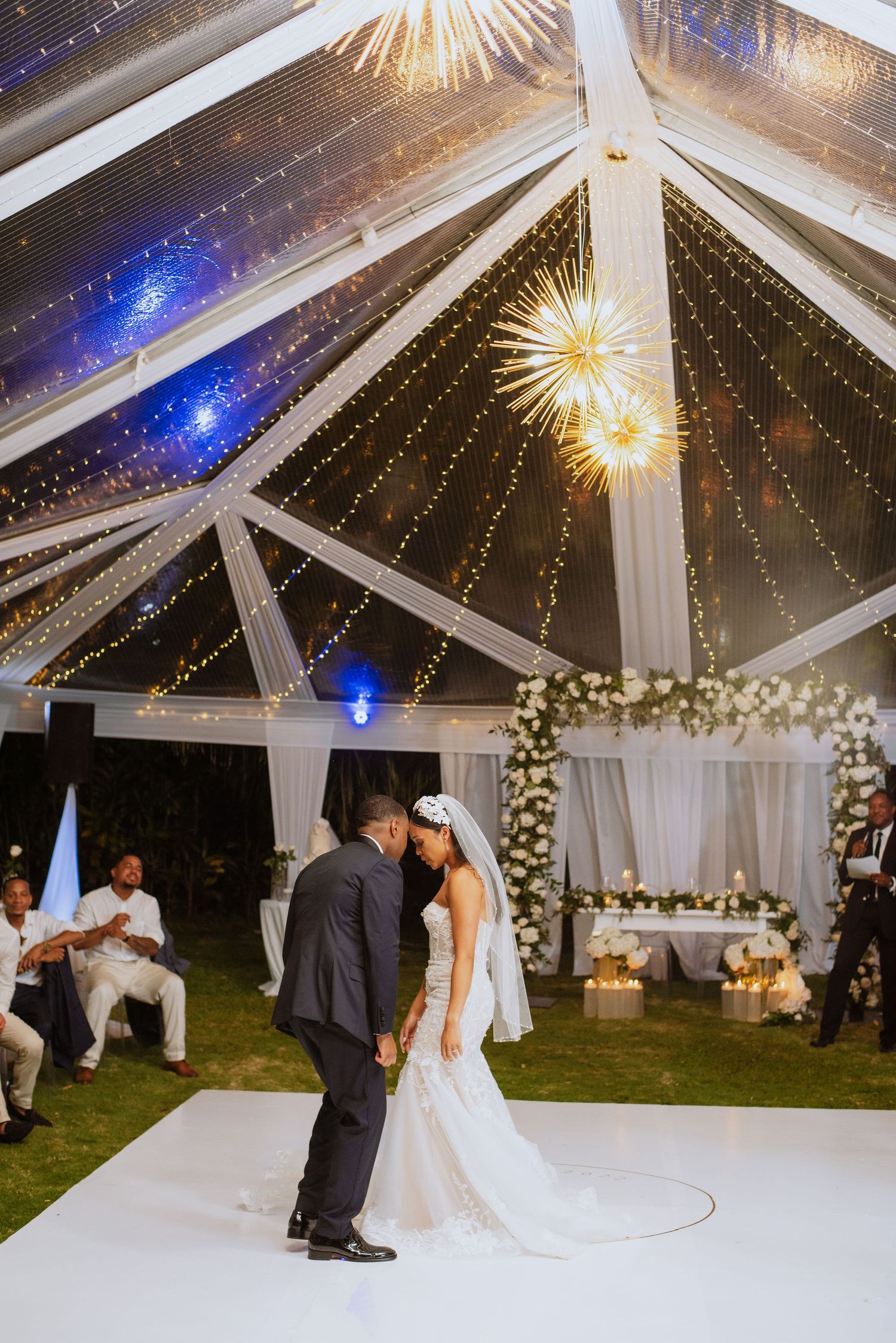 Wedding couple dances on white floor under clear tent with sparkling lights and floral arch decorations.