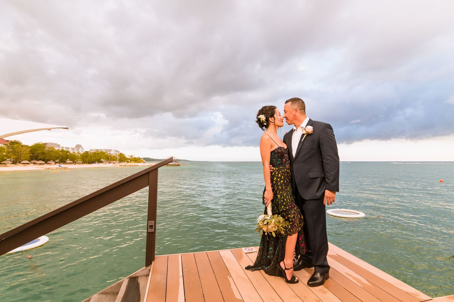 Couple shares romantic moment on wooden deck overlooking tropical beach at sunset with dramatic clouds.