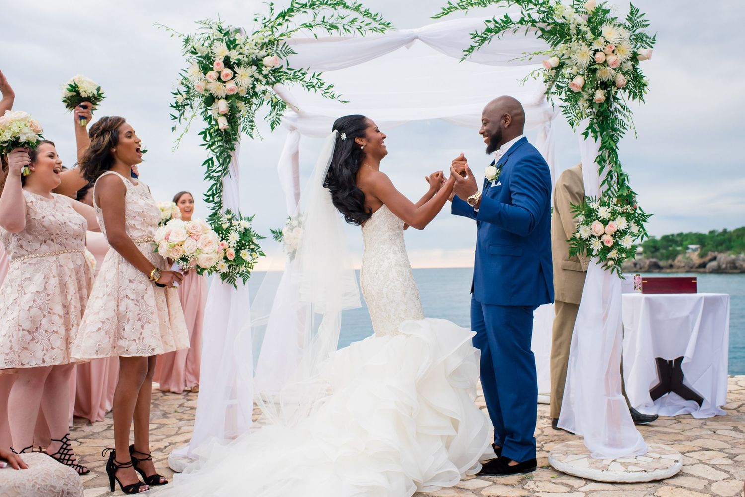 Joyful wedding celebration under a floral arch with bridesmaids in pink dresses looking on.