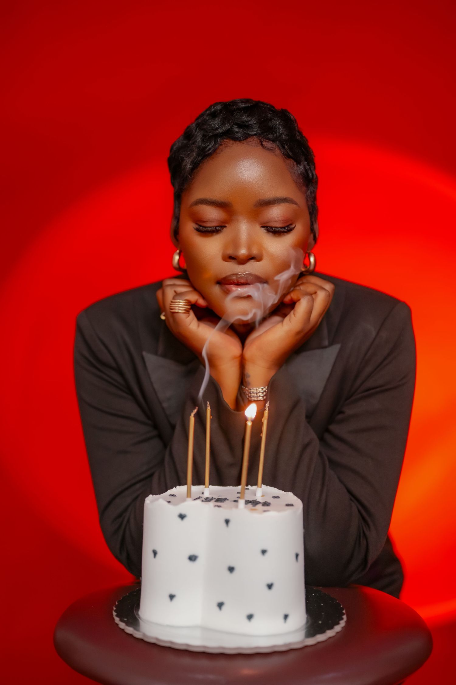 Person in black attire leaning over a white birthday cake with candles against a vibrant red background.