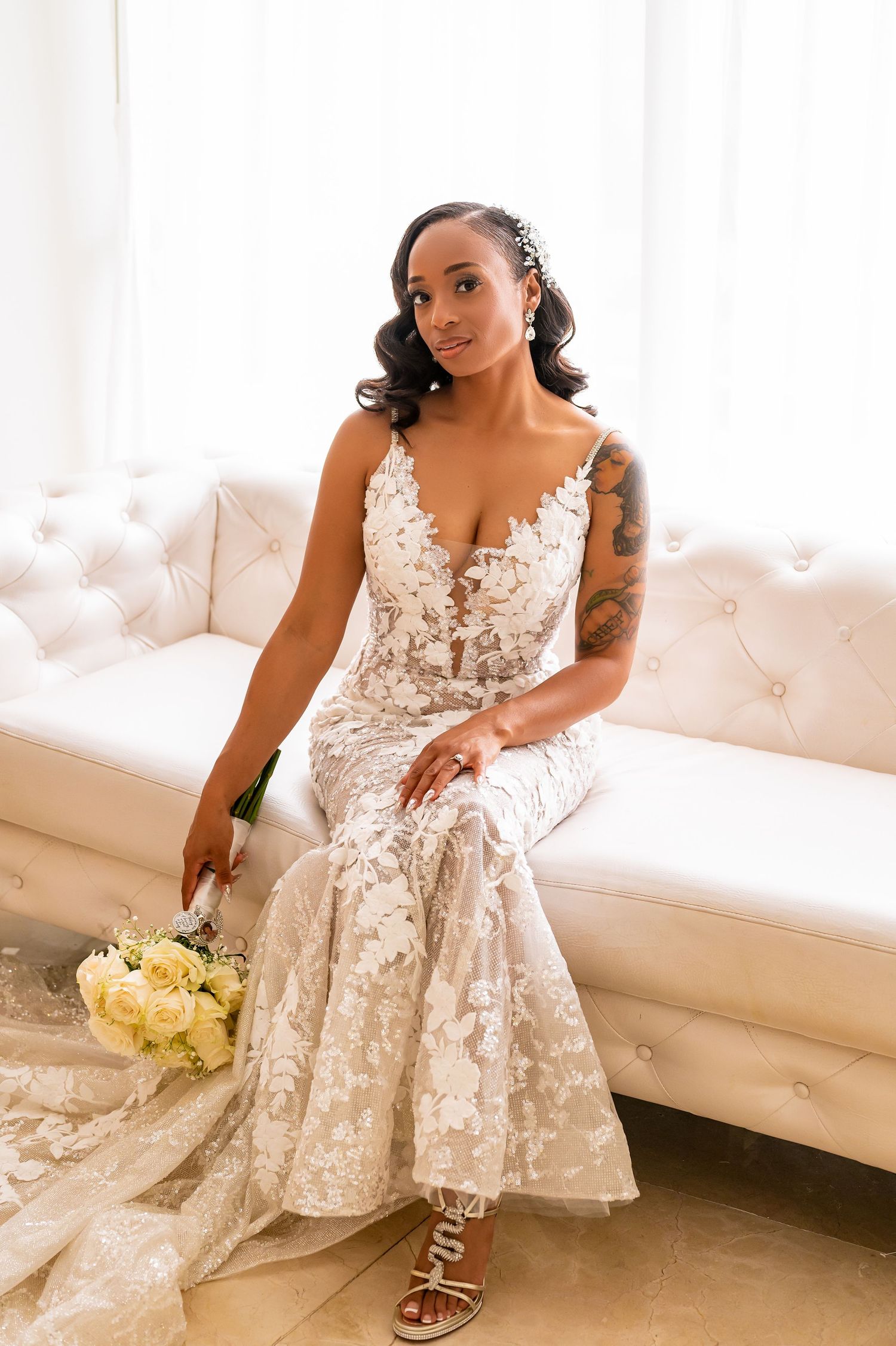 Bride in white lace dress seated on elegant tufted couch with bouquet.