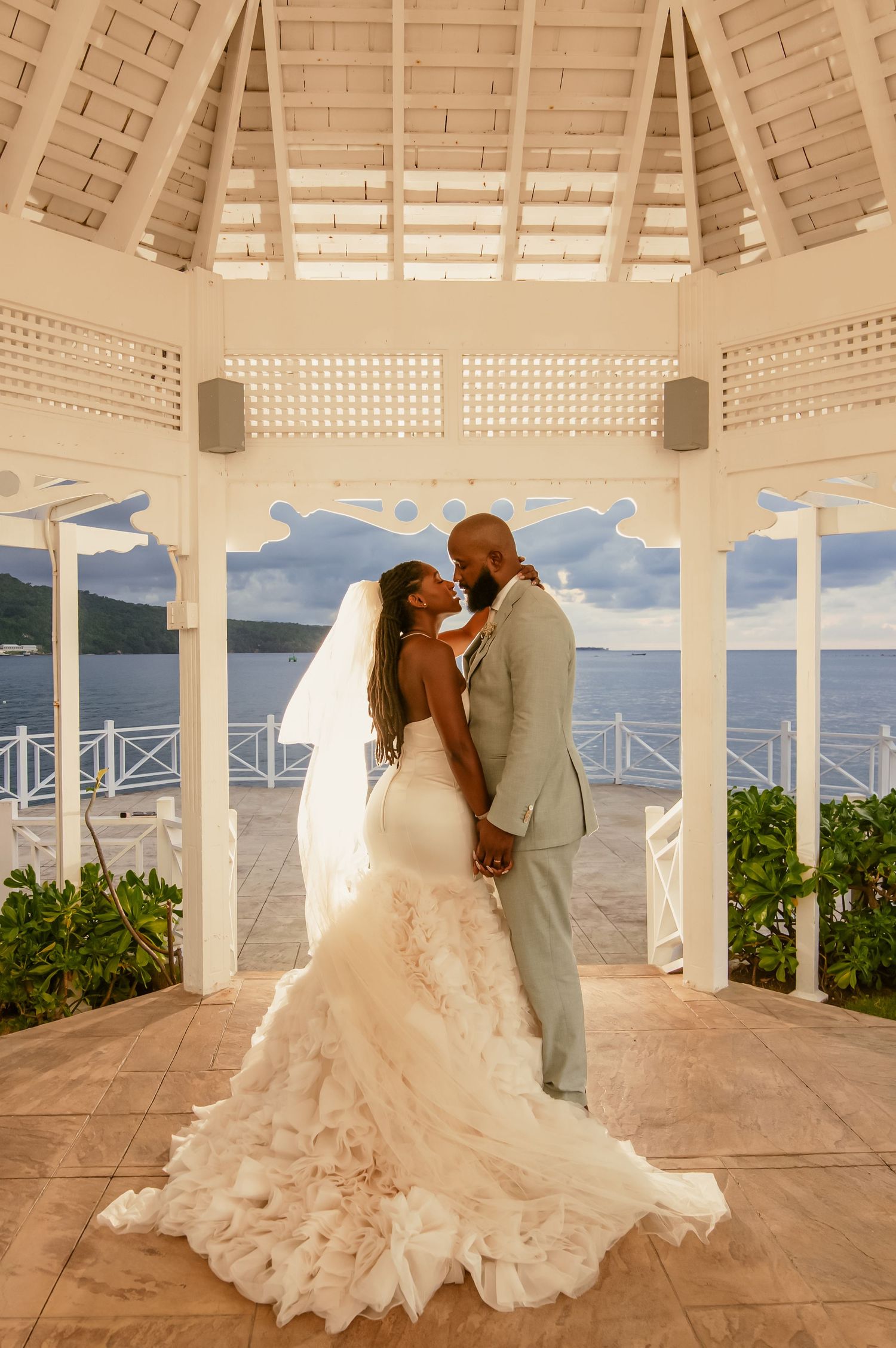 A romantic wedding couple shares intimate moments under a beachfront gazebo at sunset.