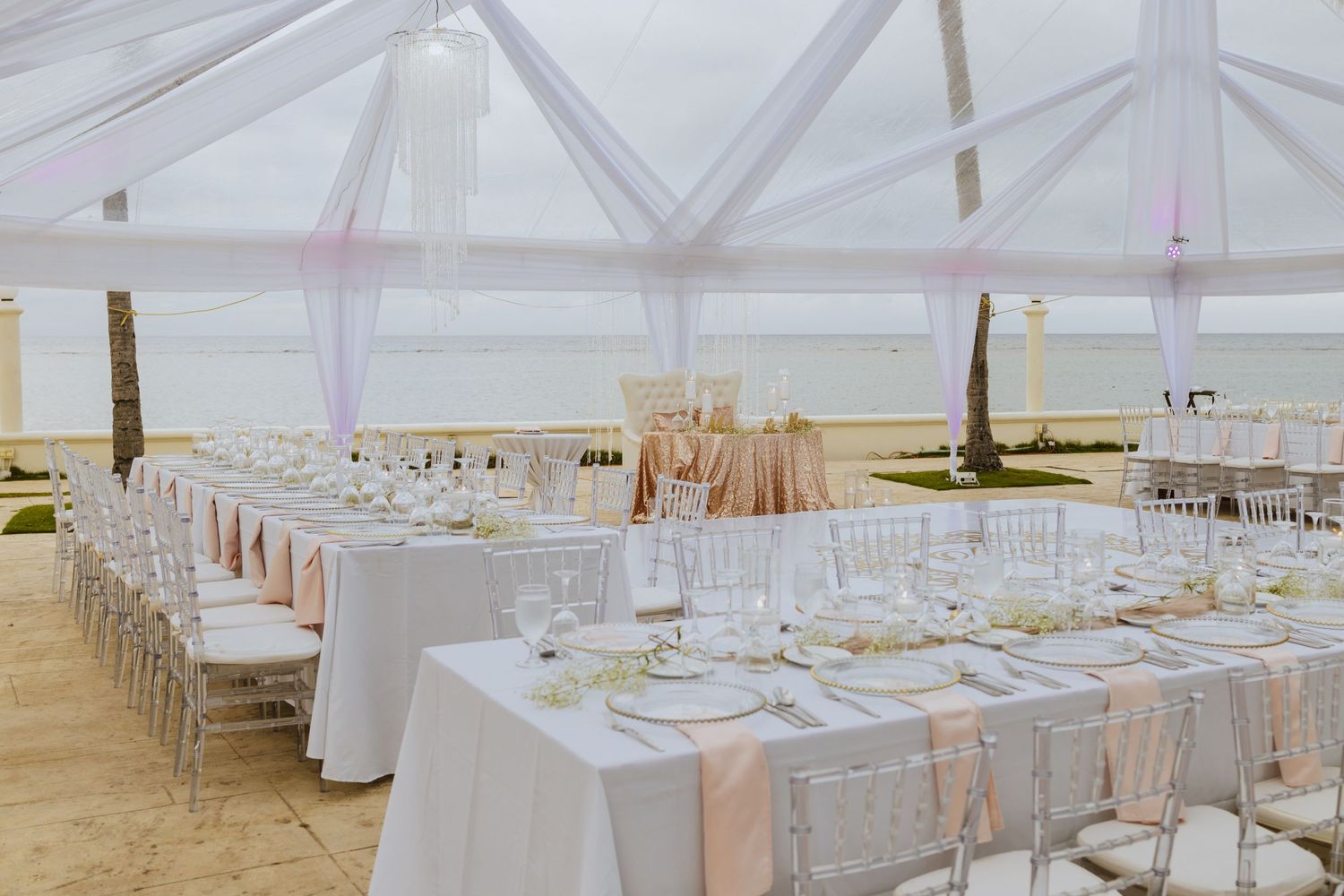 Beachfront wedding reception with white tables and chairs set under a clear tent structure.