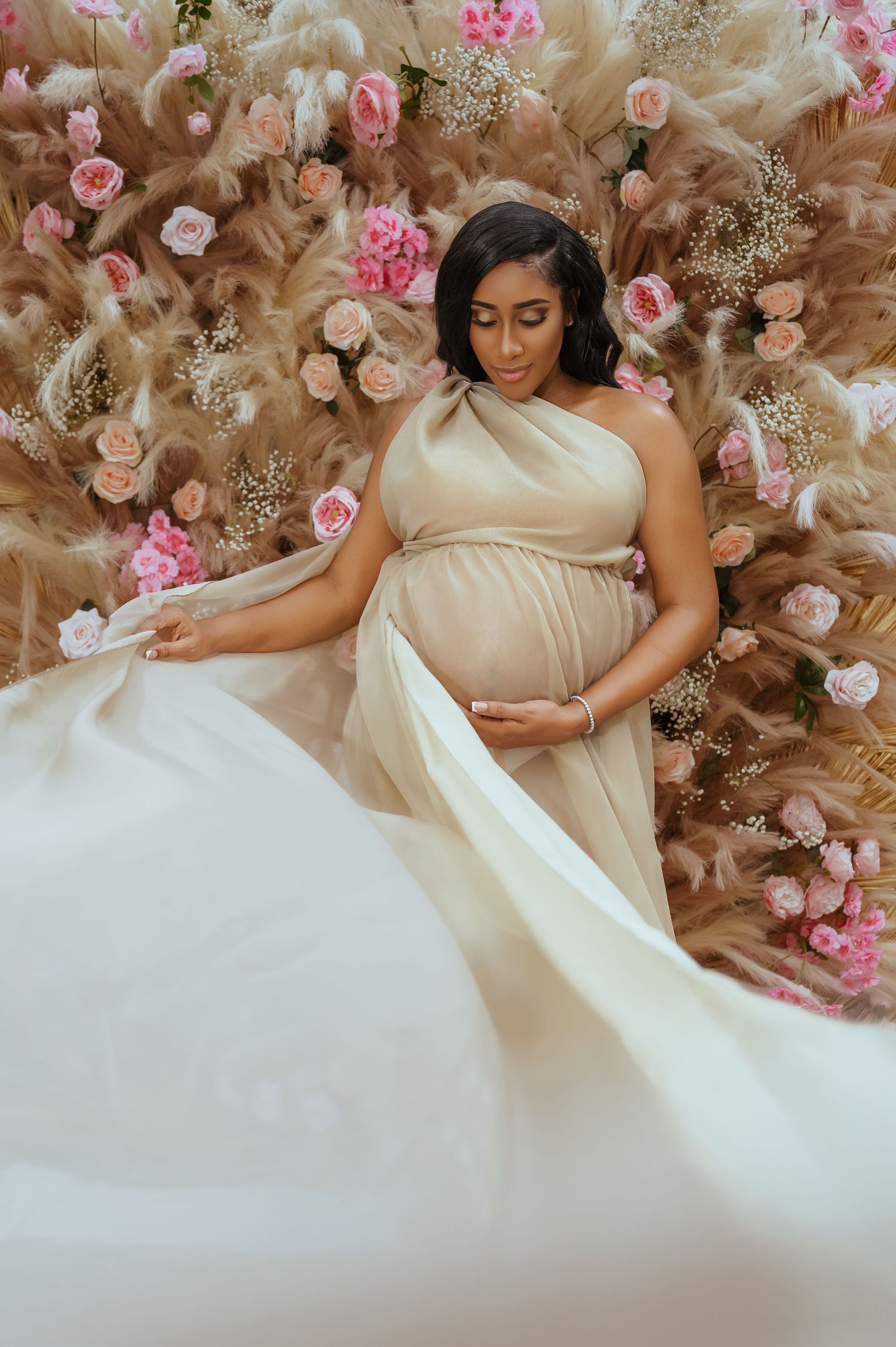 Maternity photo with pink roses and soft beige backdrop featuring flowing white dress.