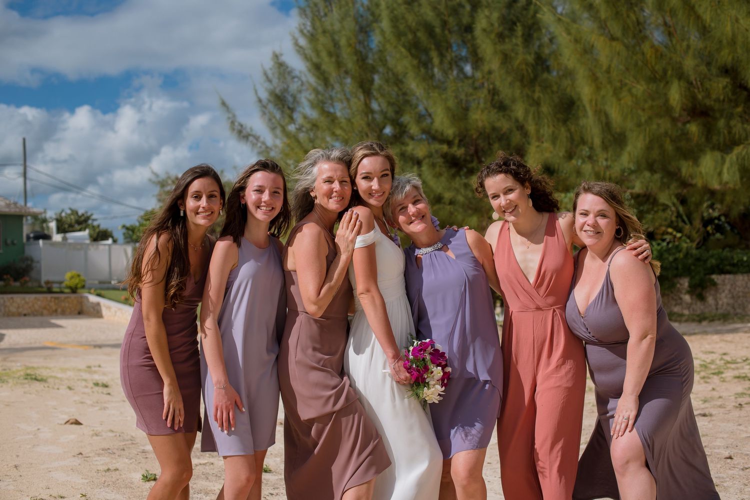 Wedding party members in coordinated purple and coral dresses stand together on a sunny beach.