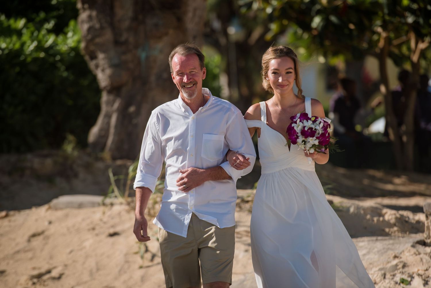 Wedding couple strolling through natural beach setting with trees.